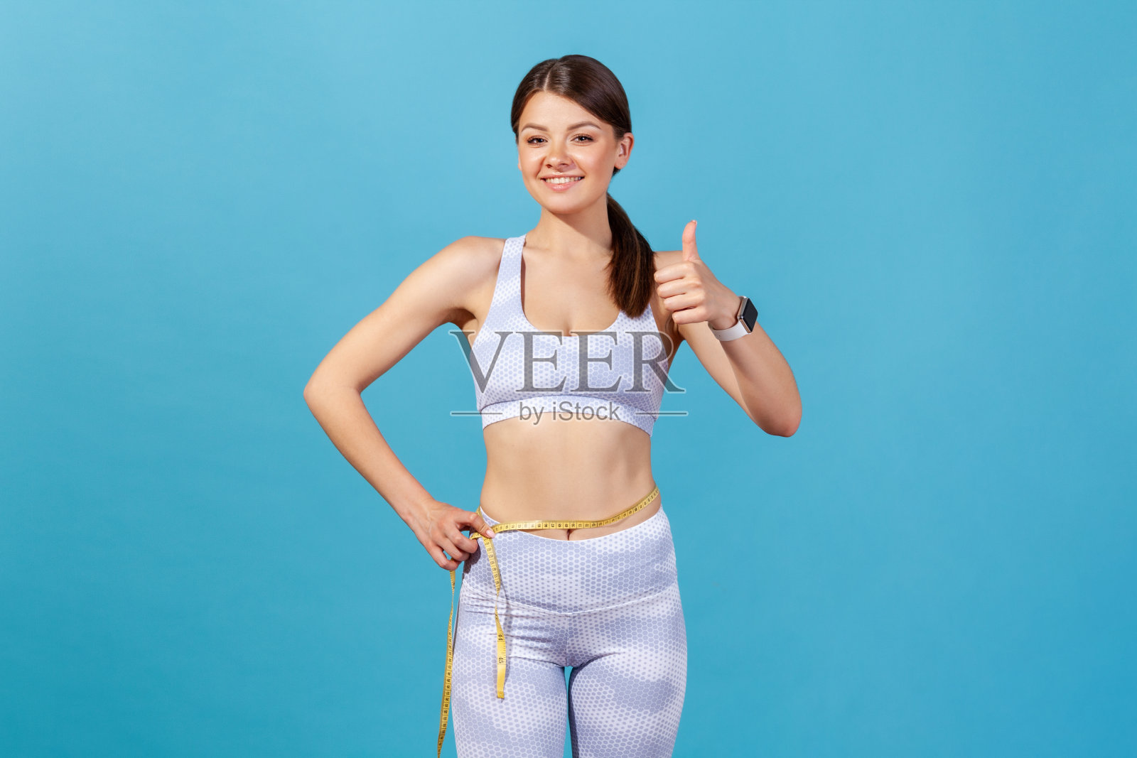 微笑积极的女人在白色运动紧身衣和顶端保持胶带线在她的腰和显示拇指向上，满意她的身体参数照片摄影图片