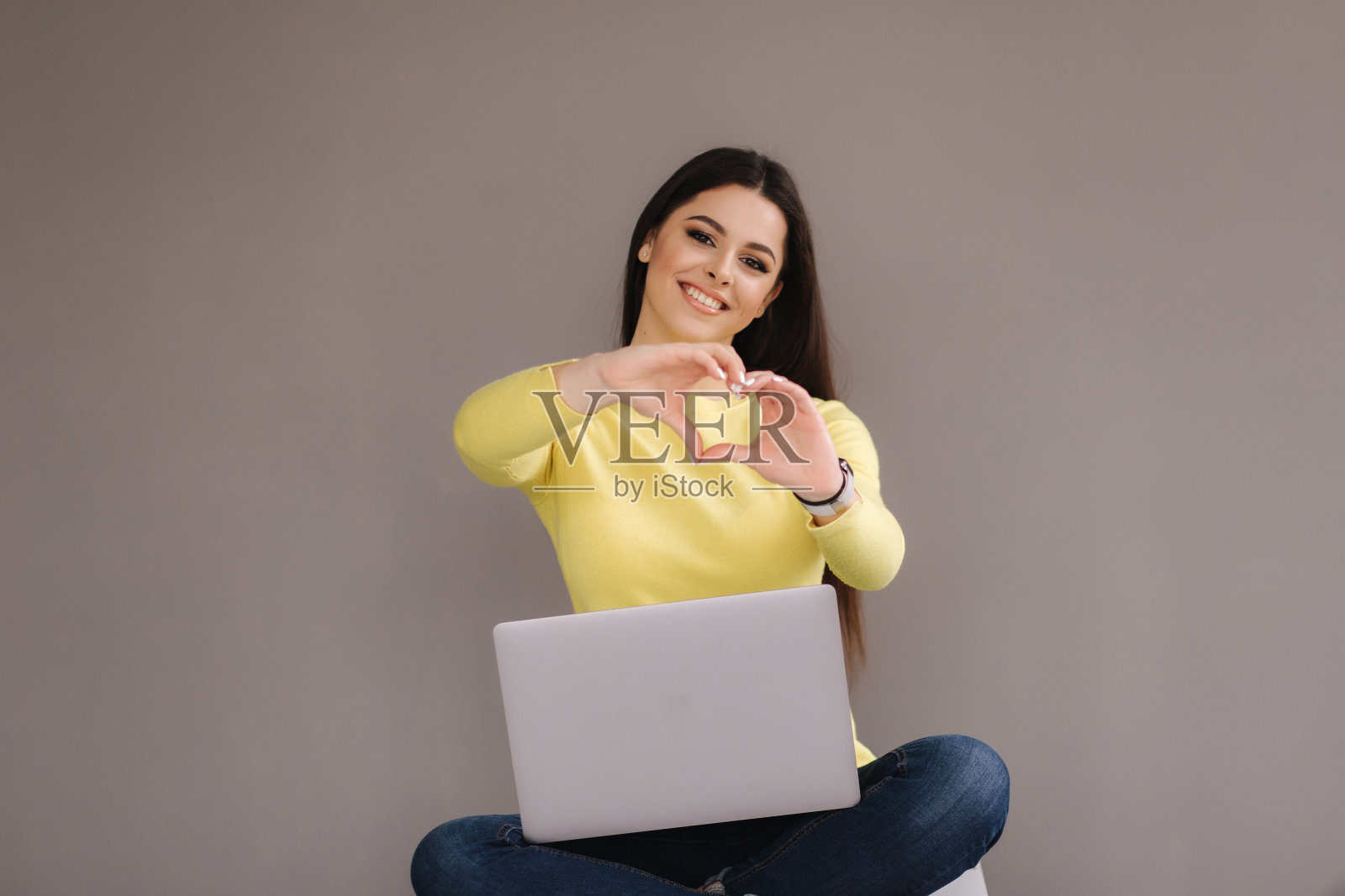 迷人的年轻女子坐在灰色背景的笔记本电脑。女人穿着黄色毛衣，用手做心形照片摄影图片