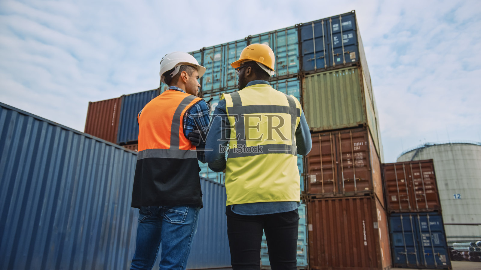 英俊的工业工程师和非裔美国监督员在安全帽和安全背心站在他们的背对着摄像机在集装箱码头。同事谈论物流运作。照片摄影图片