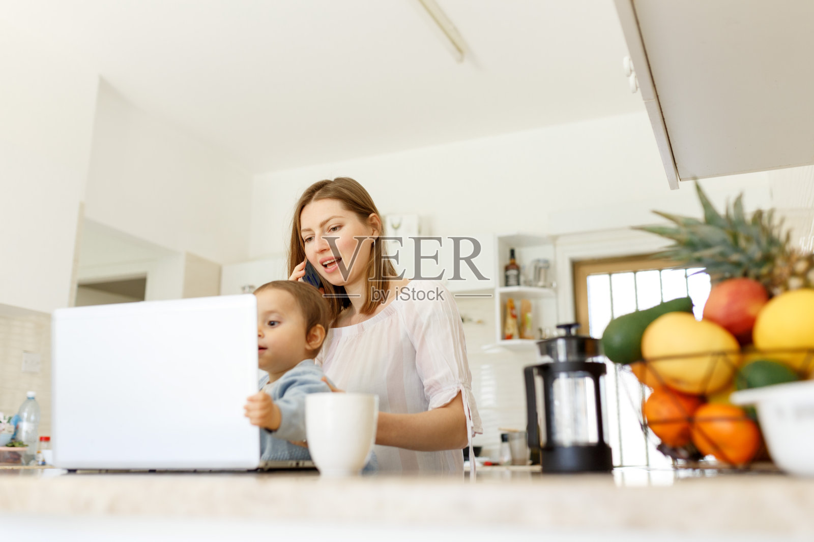 美丽的商业妈妈正在讲手机和做笔记，同时花时间与她的可爱的儿子在厨房家庭办公室。工作妈妈概念，桌上放笔记本电脑照片摄影图片