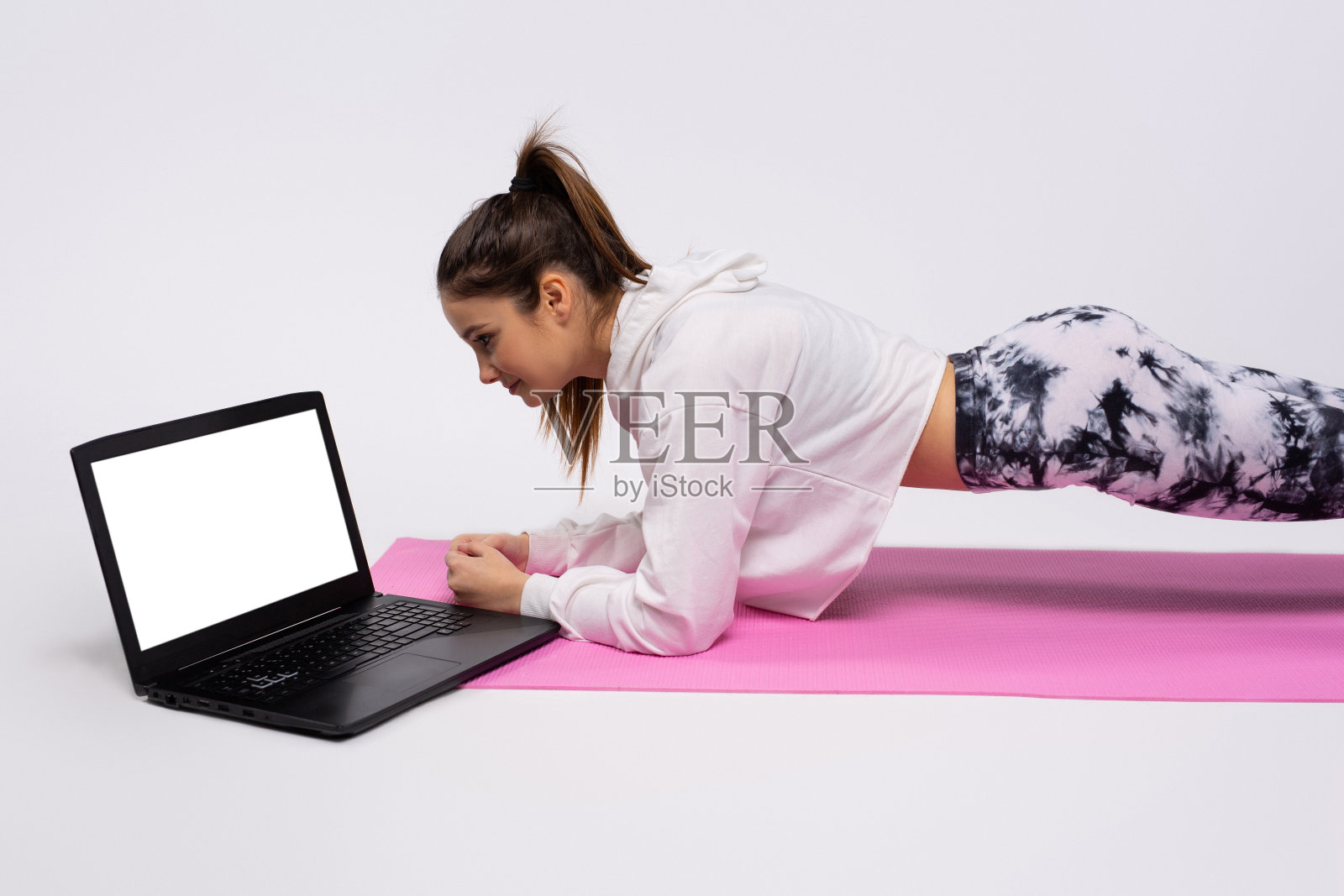 积极的女孩在运动期间穿着运动服。趴在笔记本电脑前的地毯上。白色背景。照片摄影图片