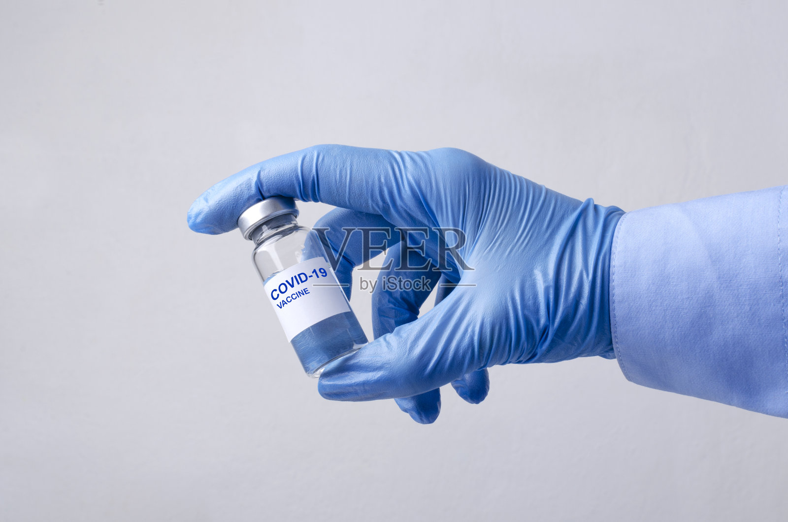 医生戴着蓝色手套，手持装有疫苗剂量的玻璃瓶，背景为灰色照片摄影图片