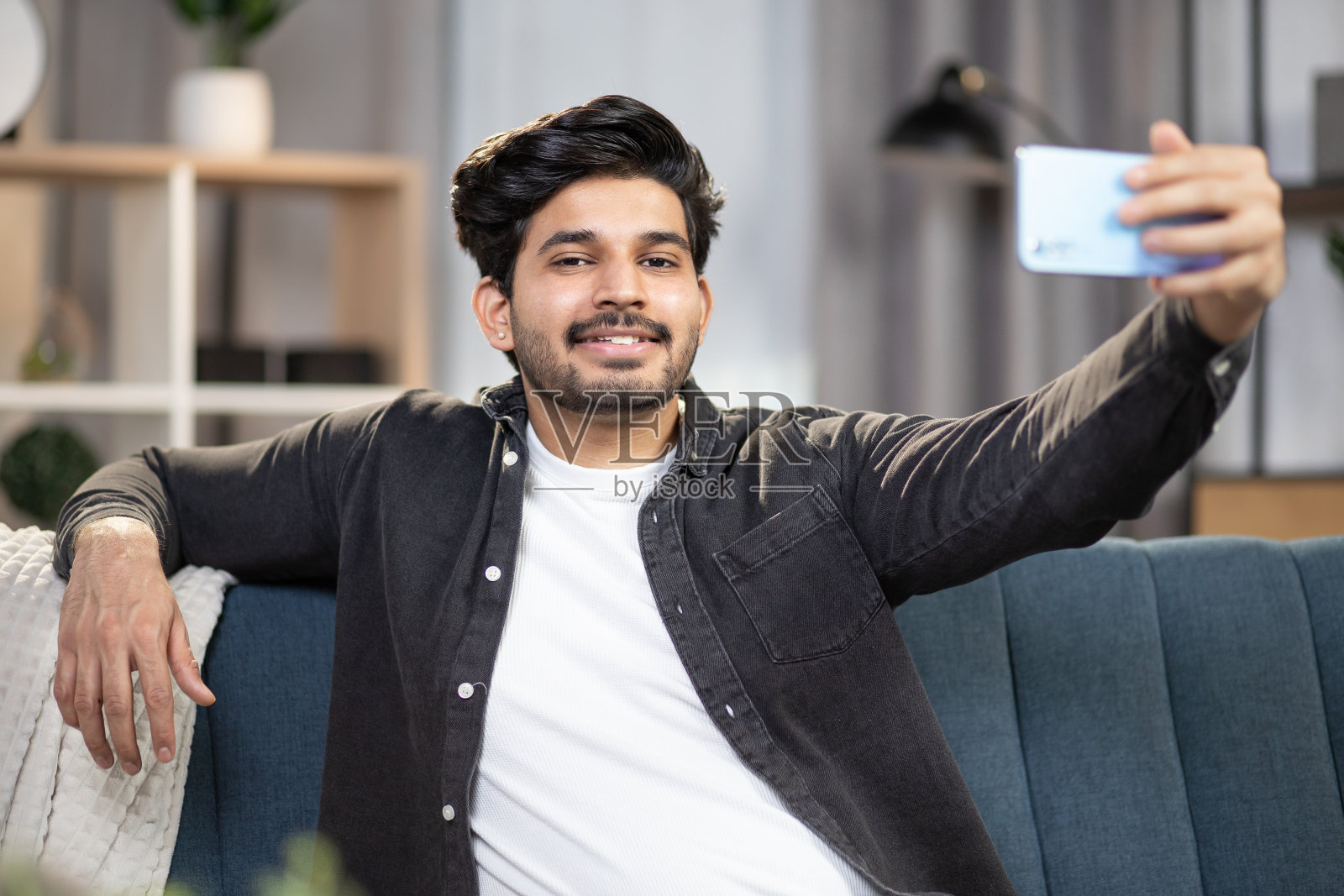 坐在舒适沙发上使用智能手机的印度英俊胡须男照片摄影图片