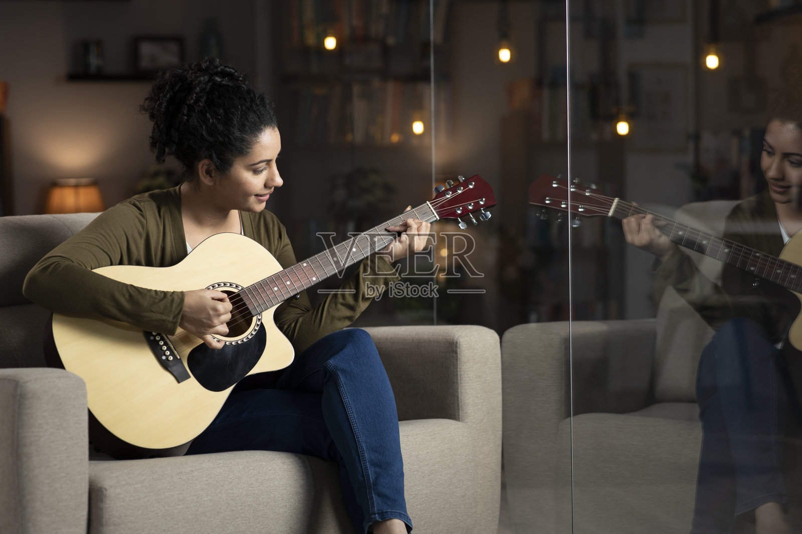 一个年轻女子坐在家里玻璃窗附近的沙发上弹吉他的照片:-库存照片照片摄影图片