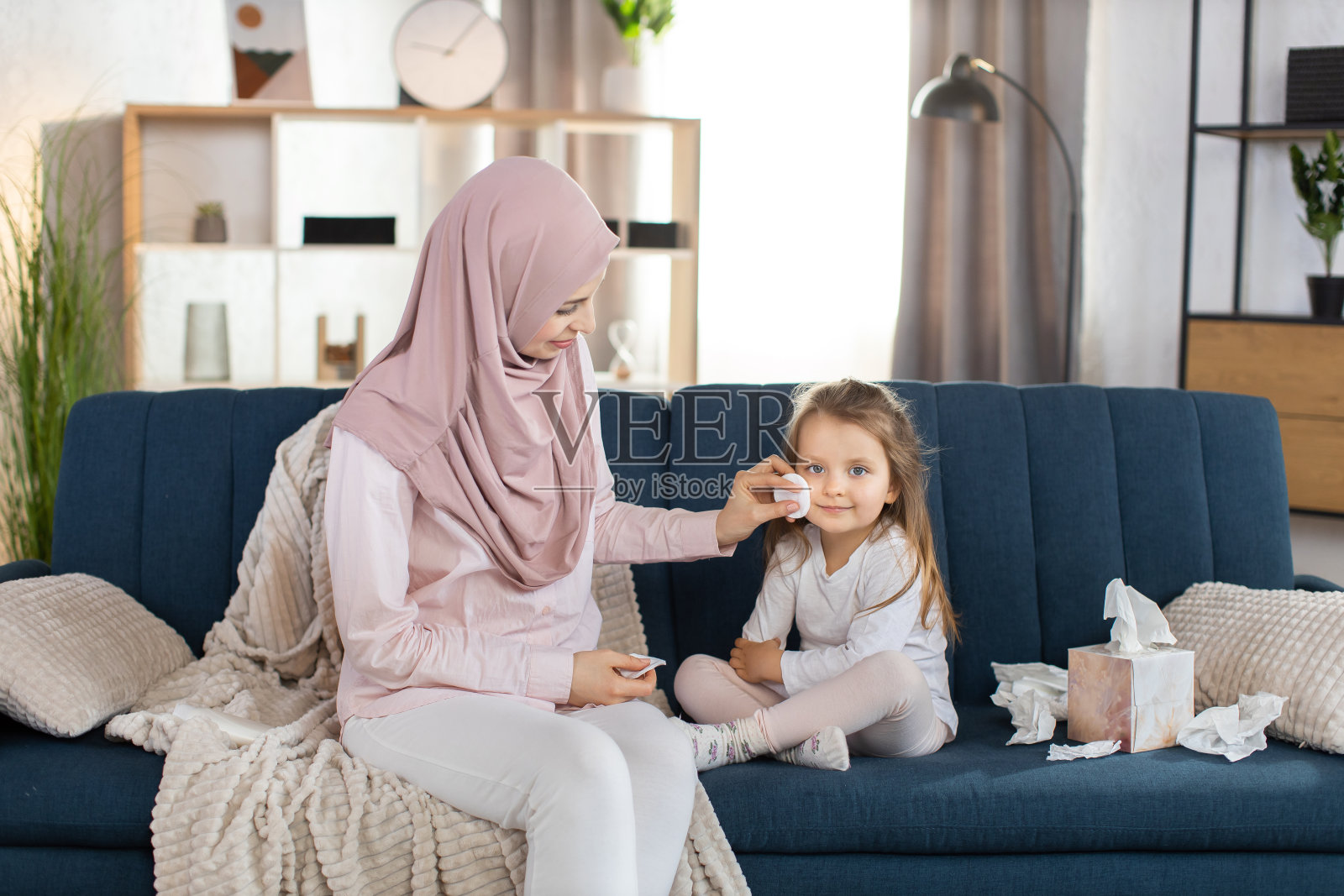 幸福的全家福。儿童保健的概念。年轻的穆斯林妇女母亲用棉花擦拭她可爱的漂亮的小女孩的脸，坐在家里的蓝色沙发上。照片摄影图片