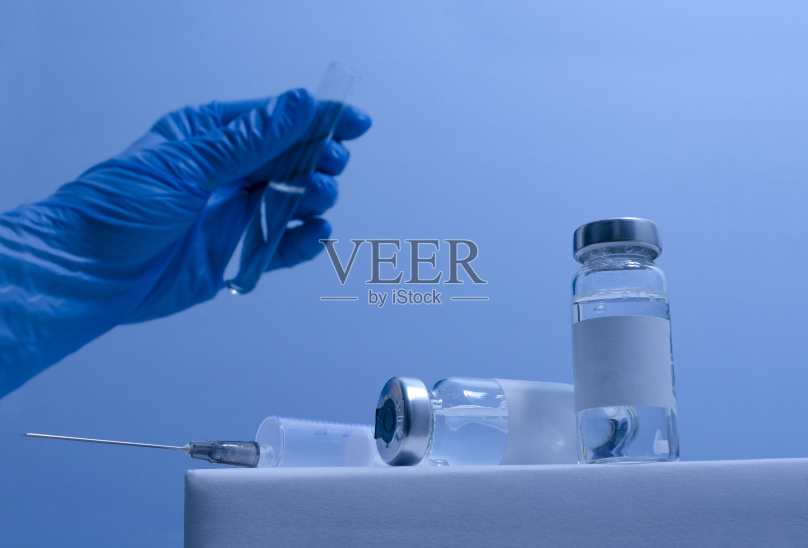 注射器的特写，玻璃瓶的疫苗与空白标签在支架上，医生的手拿实验室管在蓝色背景。致力于疫苗照片摄影图片