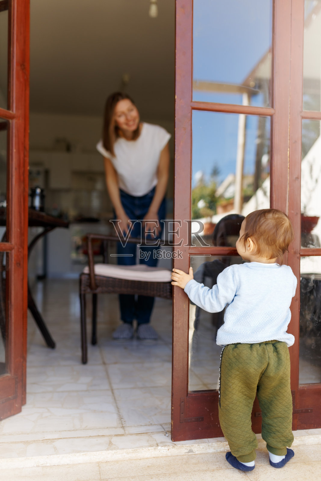 可爱的快乐的年轻女子在家里穿捉迷藏的侧面镜头。可爱可爱的婴儿宝宝从背后吮吸着奶嘴看着妈妈，玩耍着照片摄影图片