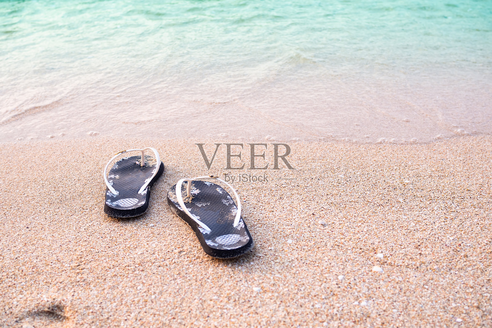 人字拖在沙滩上，柔和的波浪模糊了。黑色女式拖鞋。泰国芭堤雅的大海。夏季旅游度假概念。照片摄影图片
