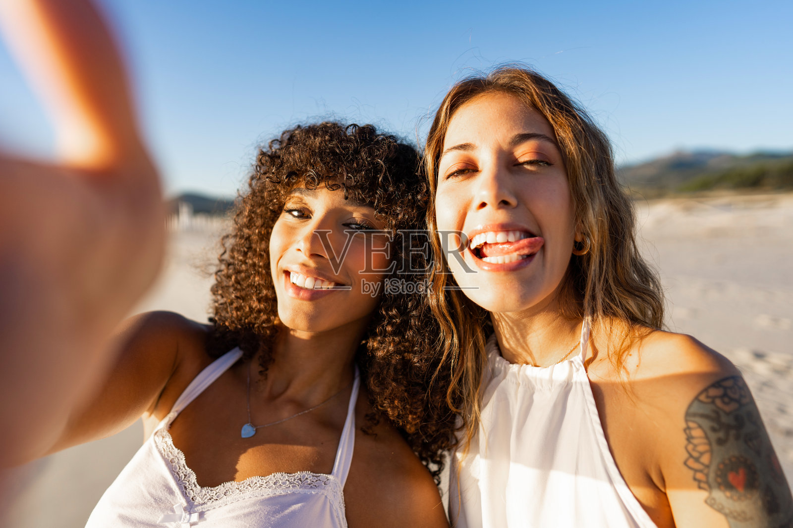 两个快乐的女人，多种族最好的朋友，穿着波西米亚衣服在海滩上自拍，滑稽的舌头脸。纹身女孩与美国黑人女友在度假时拍了一幅有趣的自画像照片摄影图片