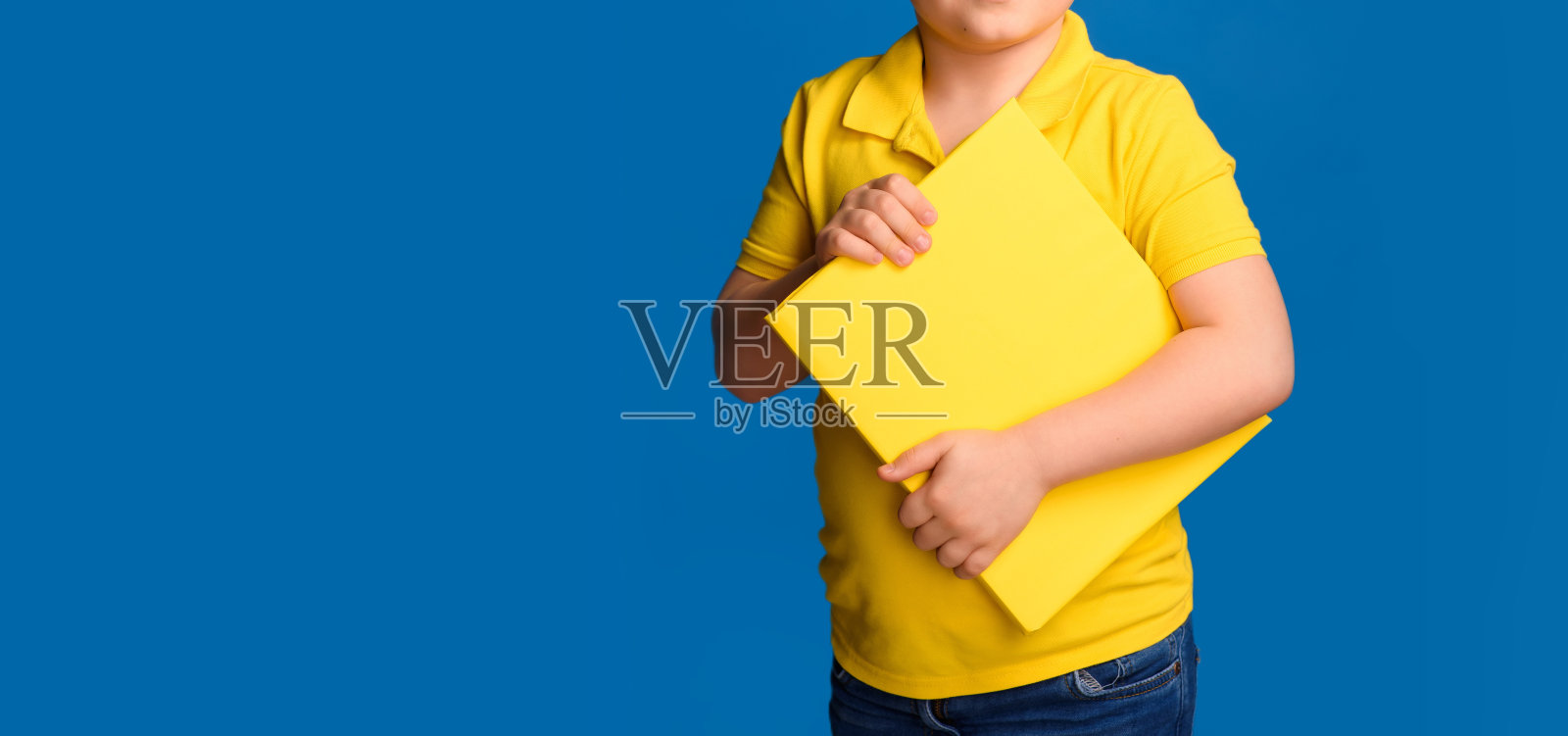一个穿着黄色t恤的小男孩拿着一本黄色的书在蓝色的背景上。照片摄影图片