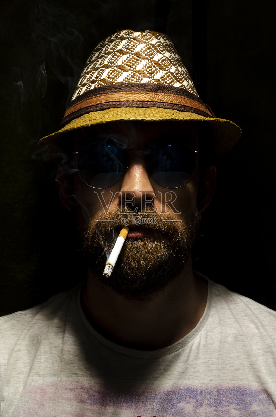 一个戴着太阳镜和帽子的大胡子男人的肖像。那人抽着烟。在黑色的背景上。照片摄影图片