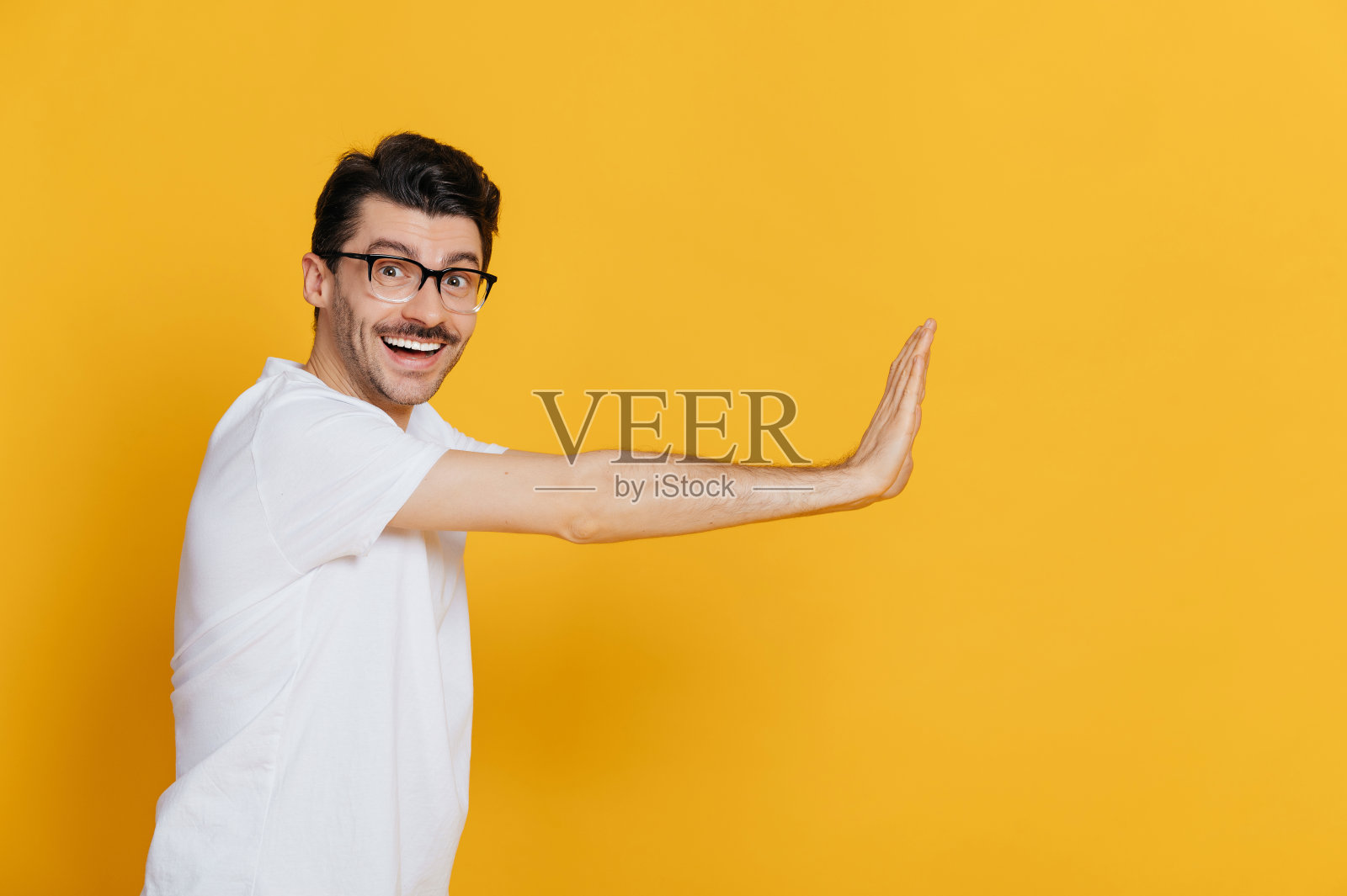 惊奇、吸引、兴奋、尖叫的白人男子戴着眼镜和白色基本的t恤，向空旷的空间手势看着相机，而站在孤立的橙色背景照片摄影图片