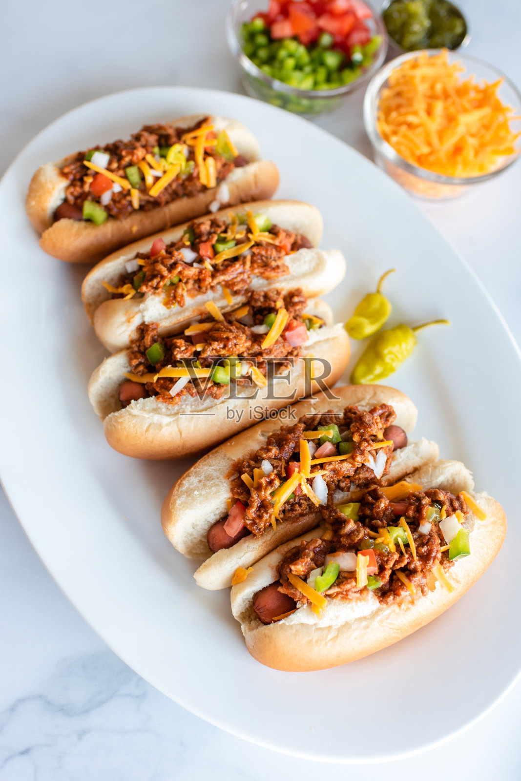 特写:白色大理石柜台上的一盘辣椒热狗和配料。照片摄影图片