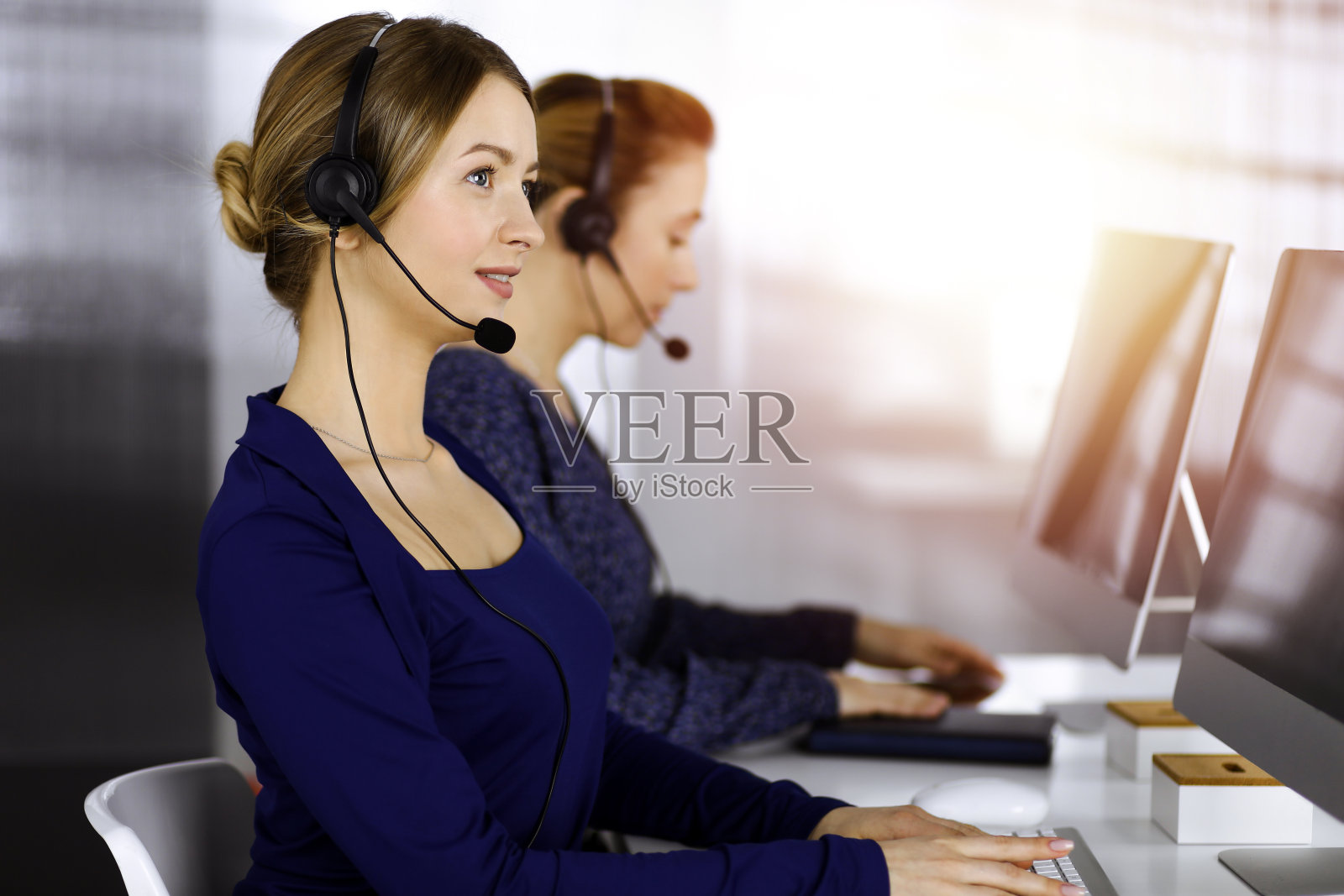 在一个阳光明媚的现代办公室里，两位女商人坐在办公桌前，用耳机与客户交谈。呼叫中心里有各种各样的人。电话营销和客户服务照片摄影图片