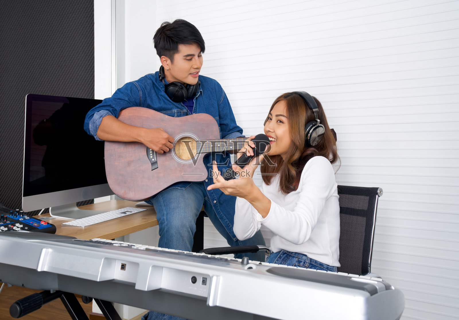 年轻的亚洲女子在白墙前唱歌。她的男朋友一起弹吉他。在专业录音棚制作音乐的音乐家。照片摄影图片