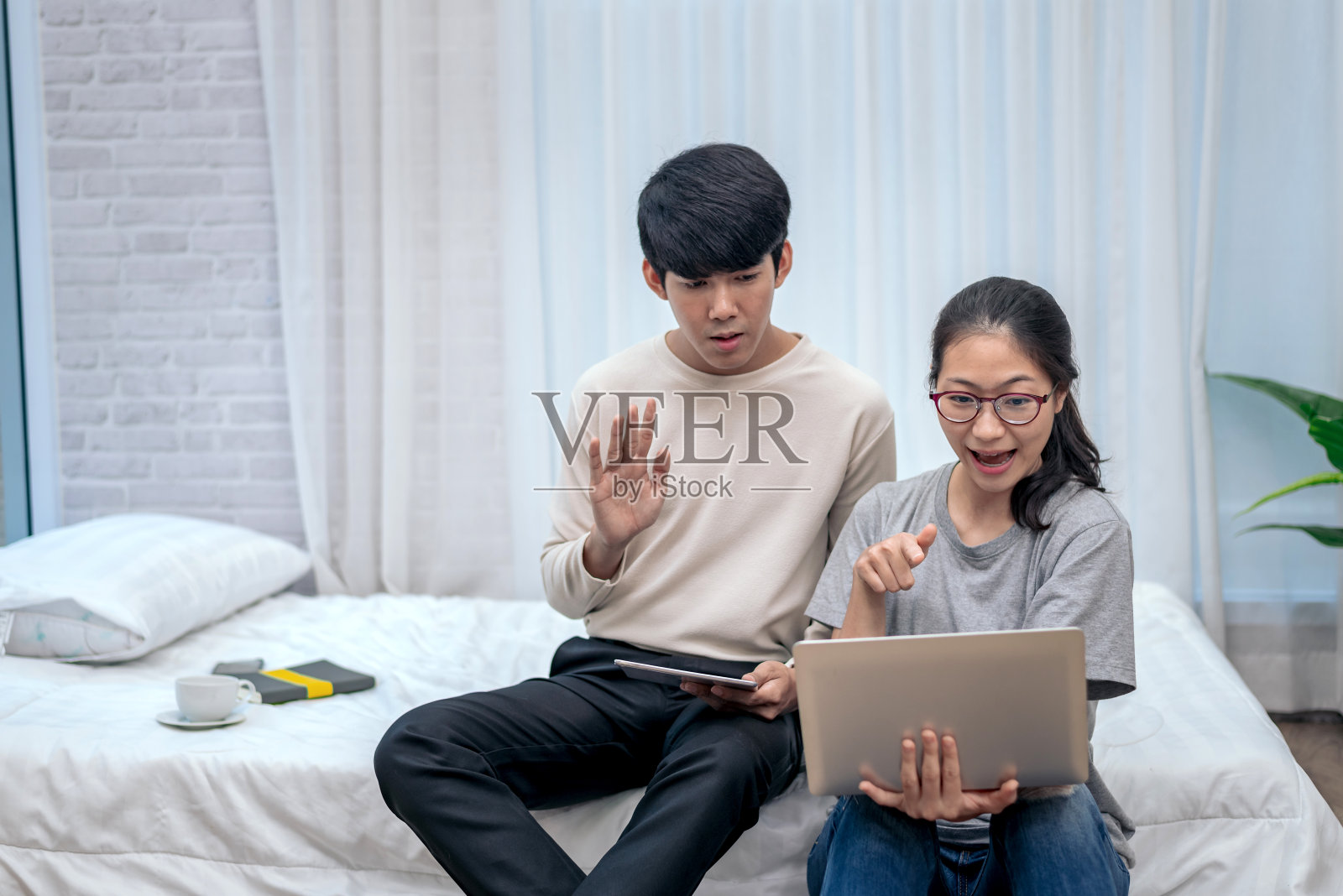 愉快的两个亚洲年轻人正在家里床上的笔记本电脑上进行视频通话。照片摄影图片