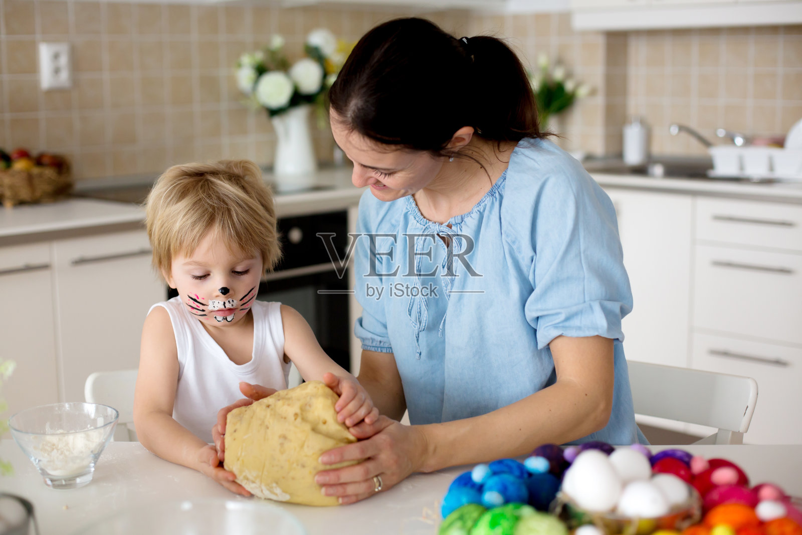 一个金发碧眼的蹒跚学步的孩子，画着兔子的脸，和年轻的妈妈在准备复活节奶油蛋糕面包，复活节甜面包的面团照片摄影图片