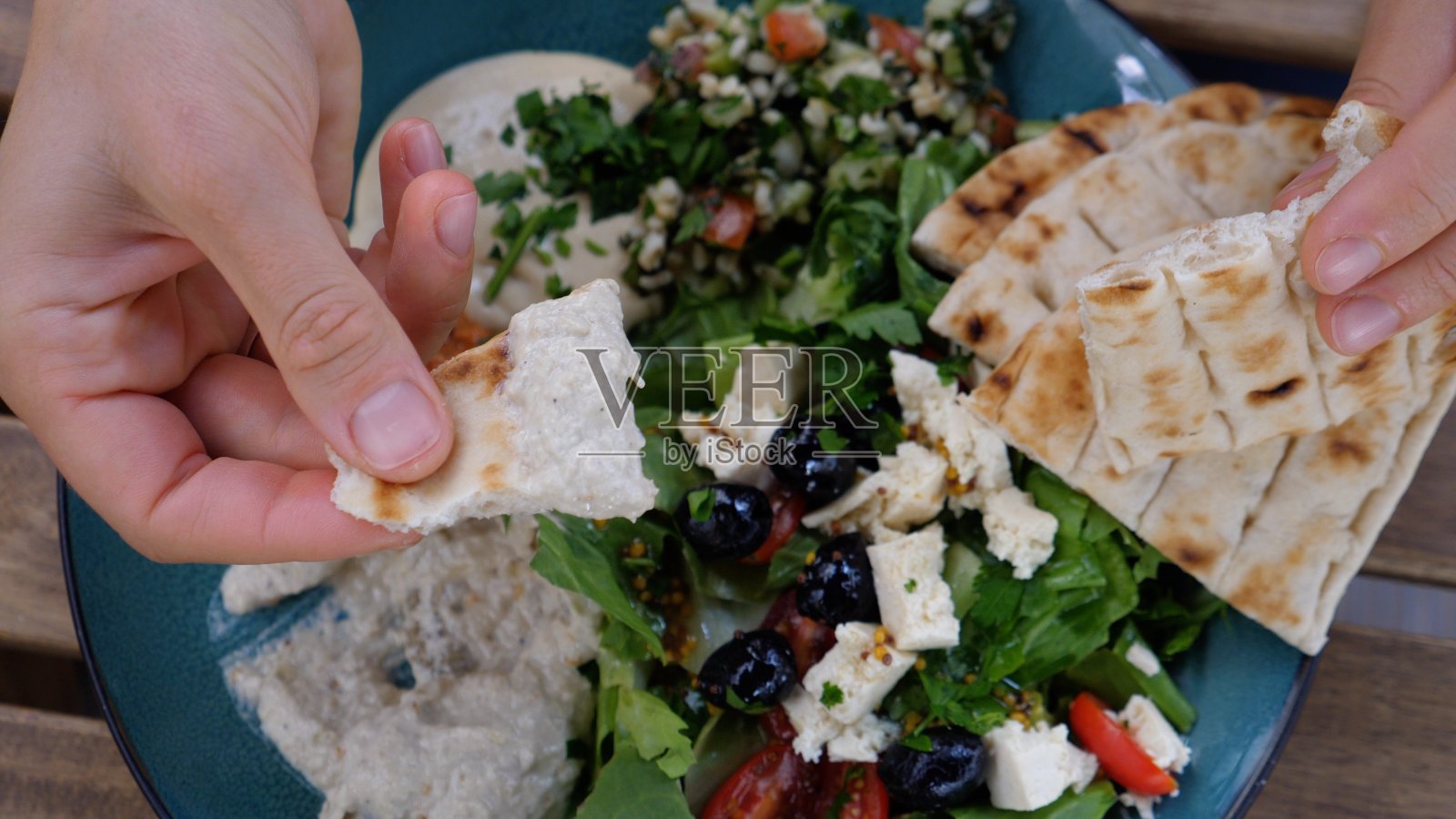手上是蘸了鹰嘴豆泥的皮塔饼。餐盘上有沙拉、蘸酱和皮塔饼。健康的中东有机烹饪照片摄影图片