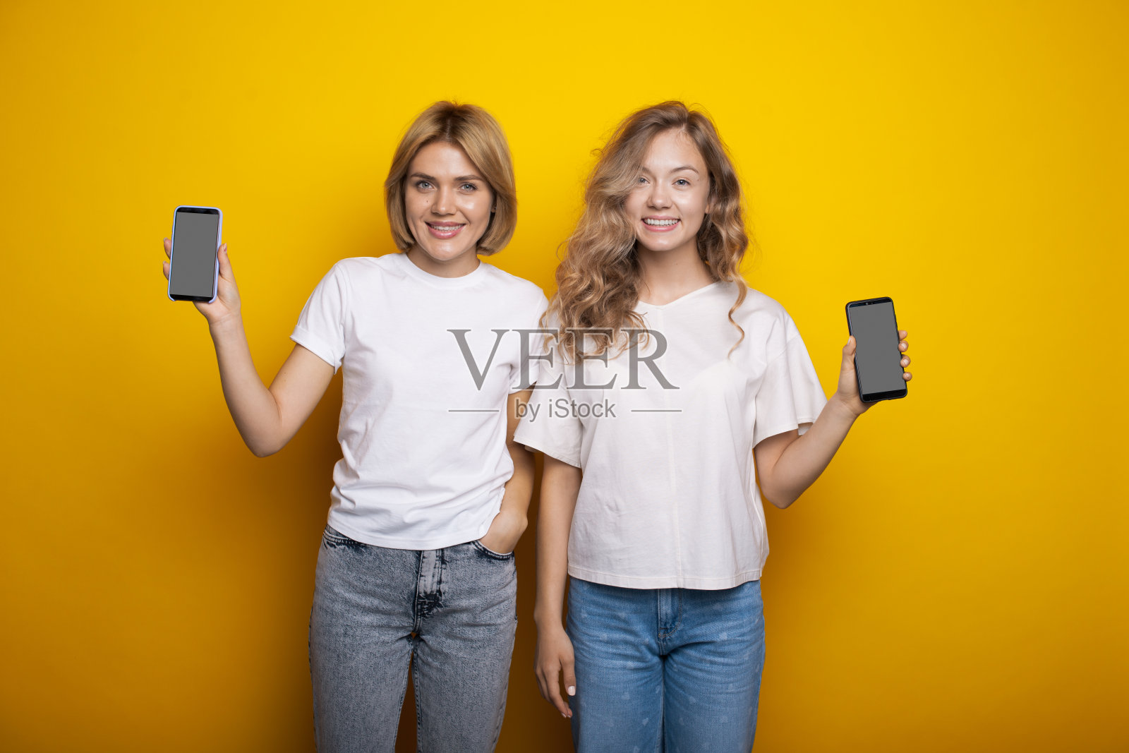 白人姐妹们正在手机屏幕上做广告，在工作室的黄色墙上微笑着照片摄影图片