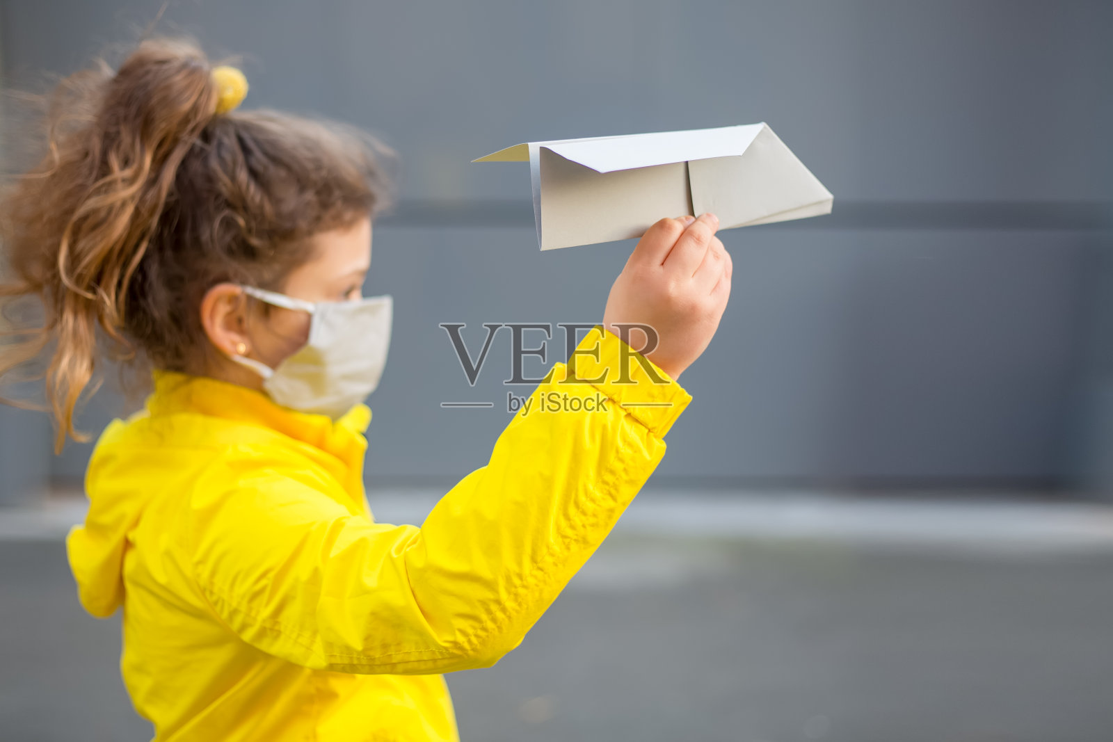 小女孩戴着灰色的防护面具，穿着黄色的夹克，在灰色的背景上拿着一架灰色的纸飞机。假日概念创意旅行covid-19流行颜色2021年照片摄影图片