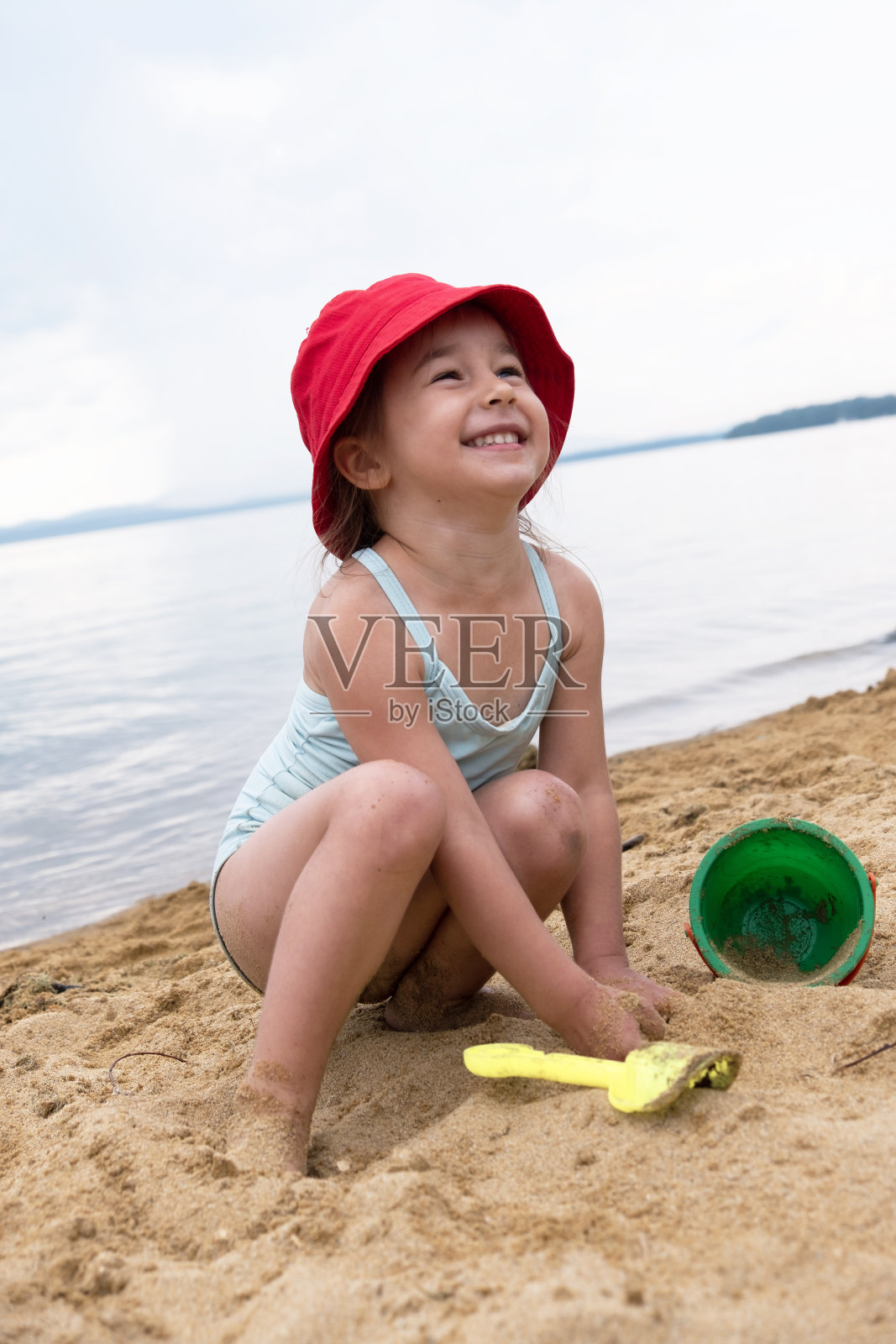 一个穿着泳衣的小女孩坐在沙滩上微笑着，拿着水桶和铲子在沙滩上玩耍，看着蓝色的天空照片摄影图片