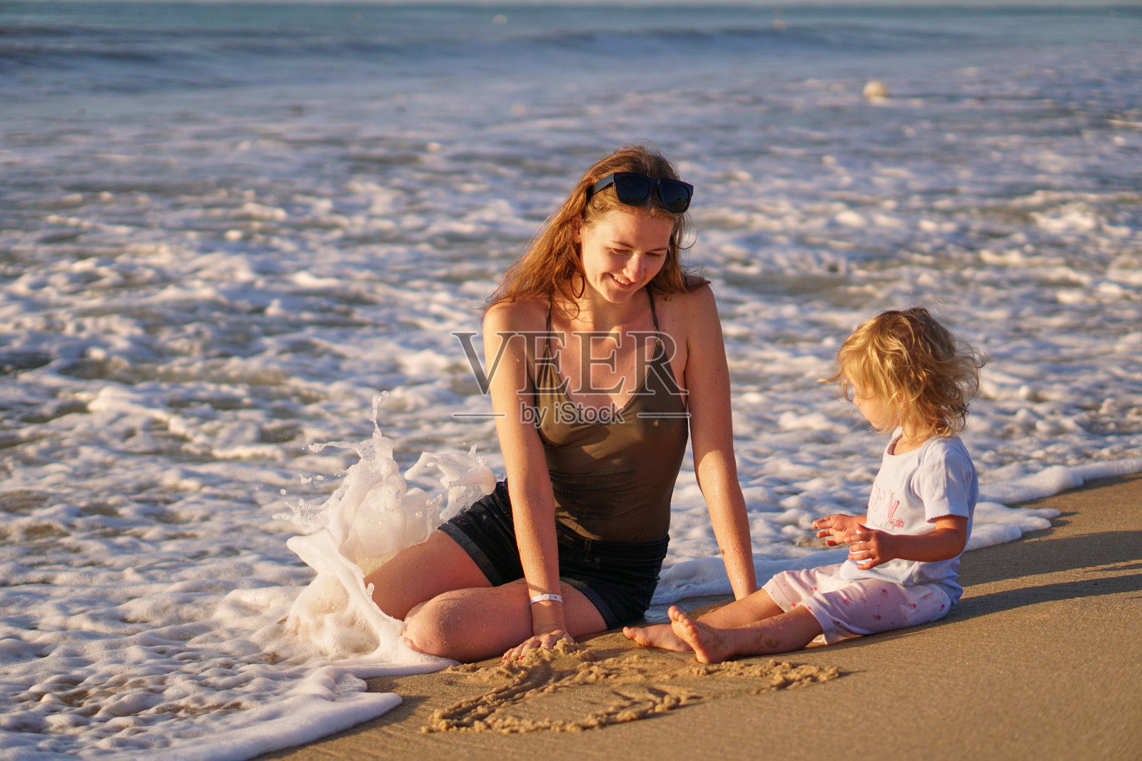 开心的笑着妈妈和女儿在海边的水里嬉笑。很高兴去度假。女孩和女人建造沙堡和海浪冲走。水覆盖了家庭。晚上玩得开心。照片摄影图片