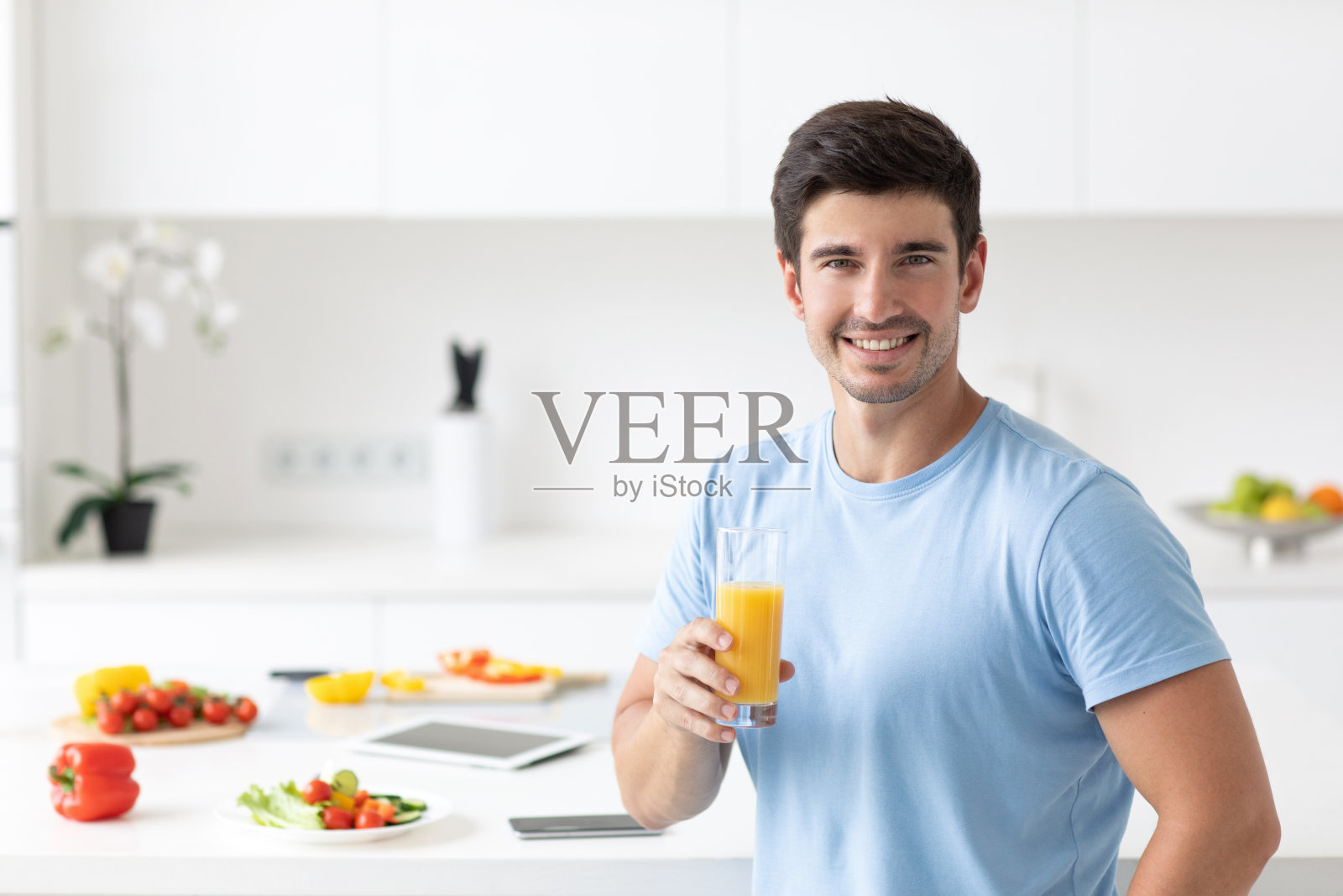 穿着蓝色t恤的帅哥手里拿着一杯橙汁在厨房里照片摄影图片