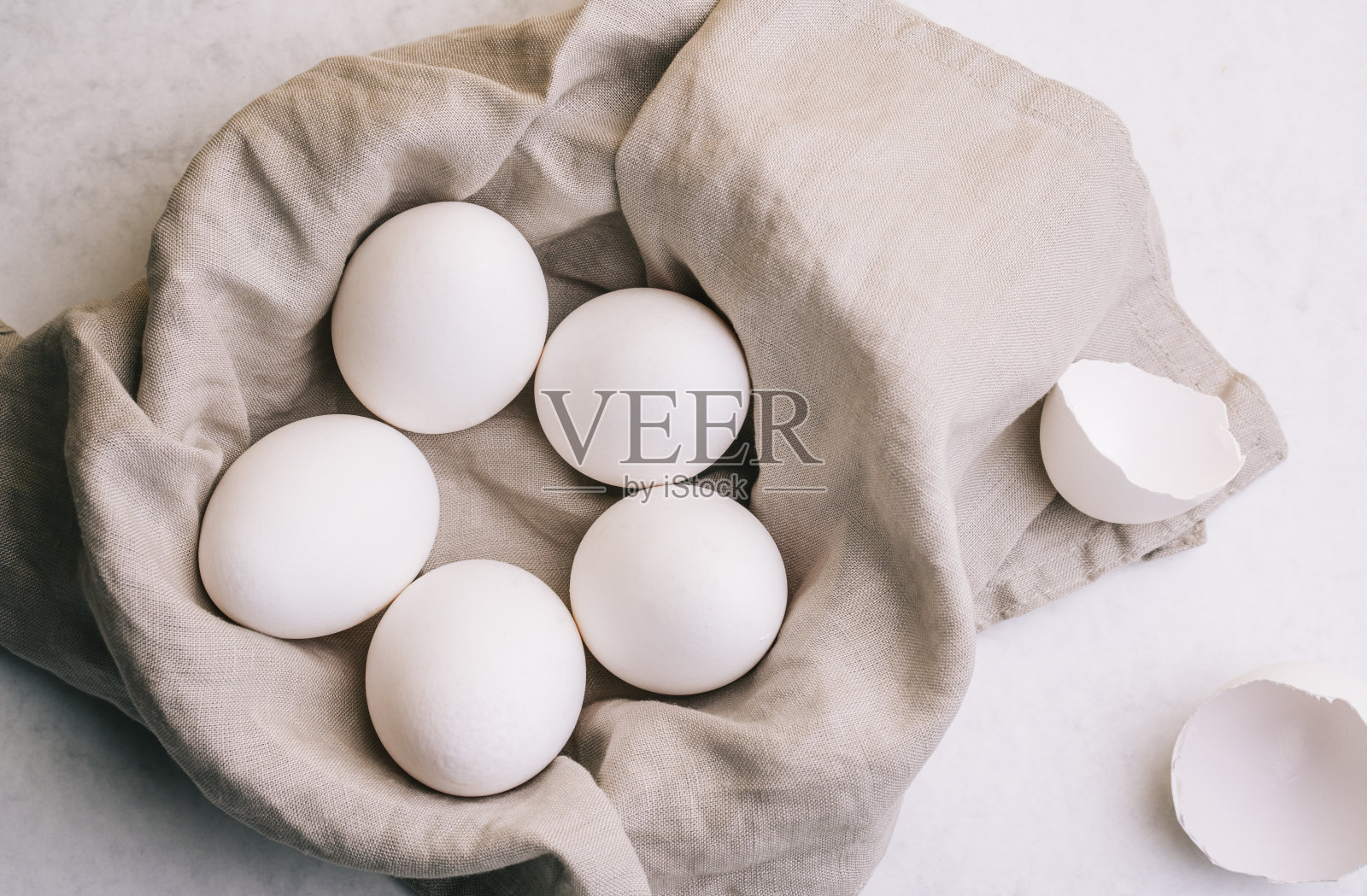一篮子白鸡蛋放在灰色的亚麻布上，旁边有两个蛋壳，背景是白色的照片摄影图片