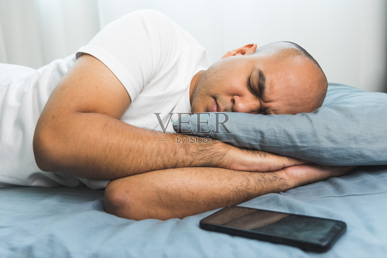 一位30岁左右的亚洲秃头男子穿着白色t恤，侧身躺在蓝色床垫和枕头上睡觉。他在家里一直睡到天亮。把智能手机放在床上照片摄影图片