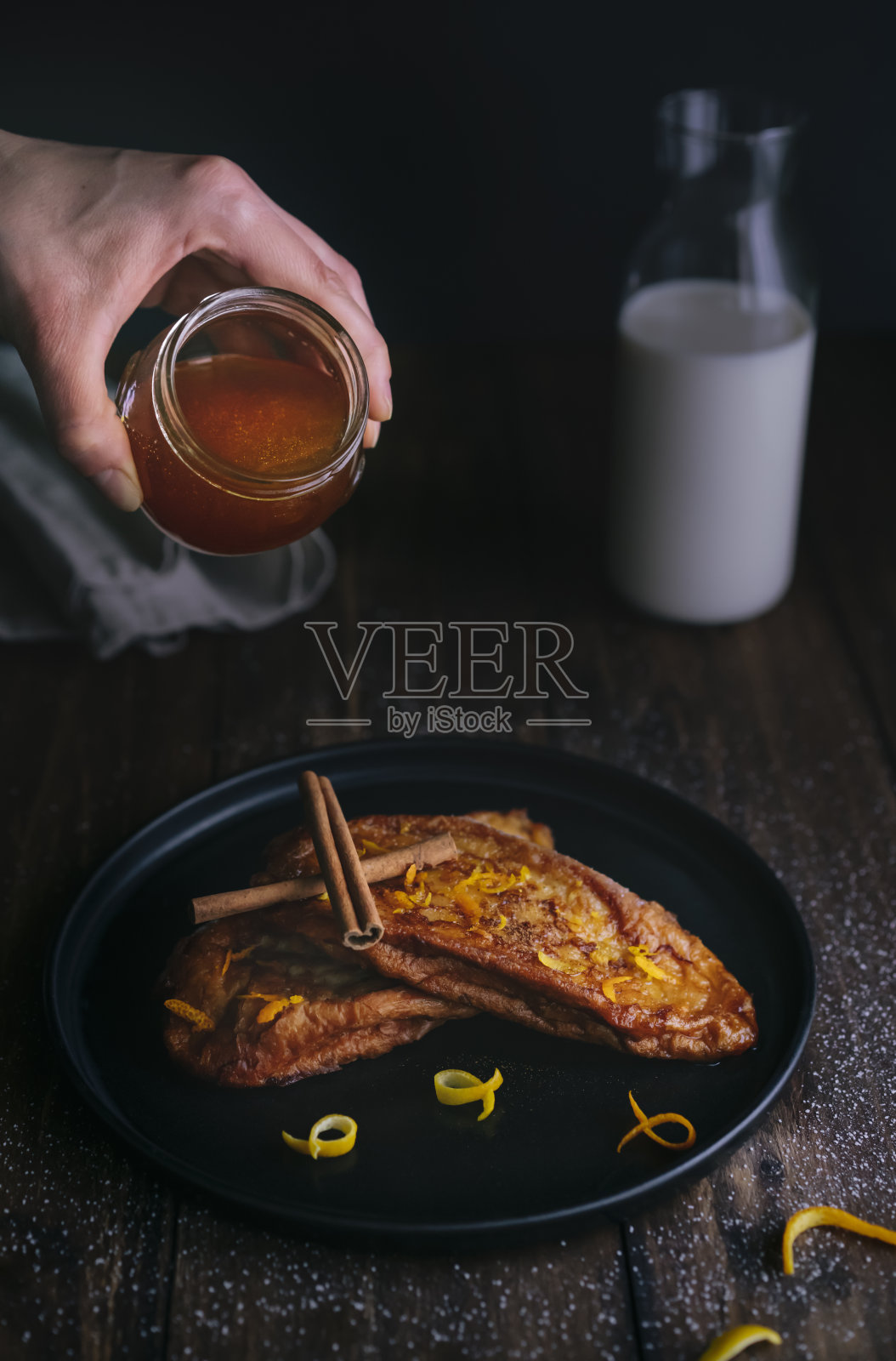 将蜂蜜倒在配有肉桂条、橙子和柠檬皮的油炸面包片上，放在一个黑色的盘子里照片摄影图片