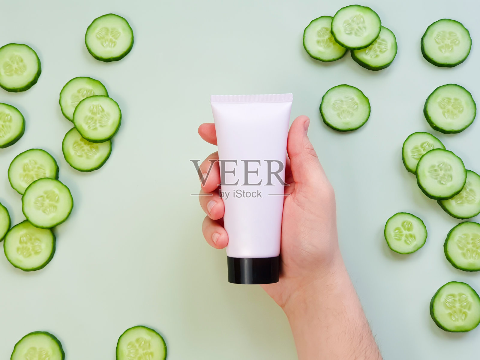男性手拿塑料无商标的粉红色奶油管与黑色盖。瓶商标和标签和新鲜黄瓜切片在绿色背景。天然有机水疗化妆品。护肤品、广告照片摄影图片