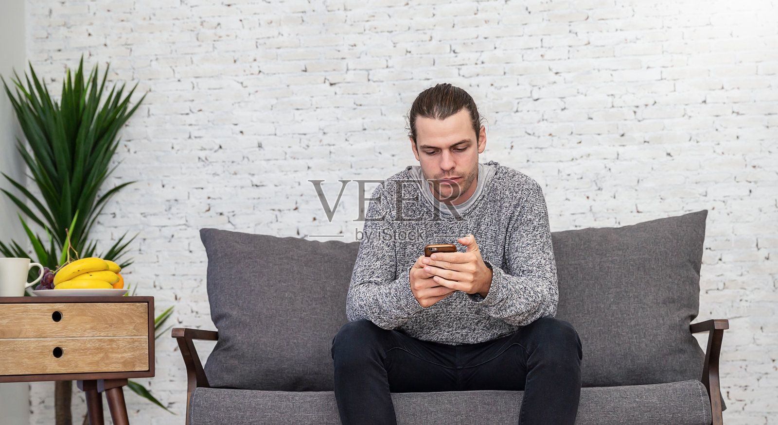 一名年轻的白人男子坐在沙发上用智能手机发短信。假期的早晨在家里的客厅放松。生活幸福平安冬天，科技连接hygge概念旗帜照片摄影图片