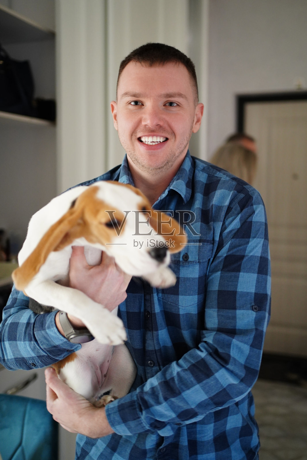 一个穿着蓝色格子衬衫的男人抱着一只比格犬。照片摄影图片