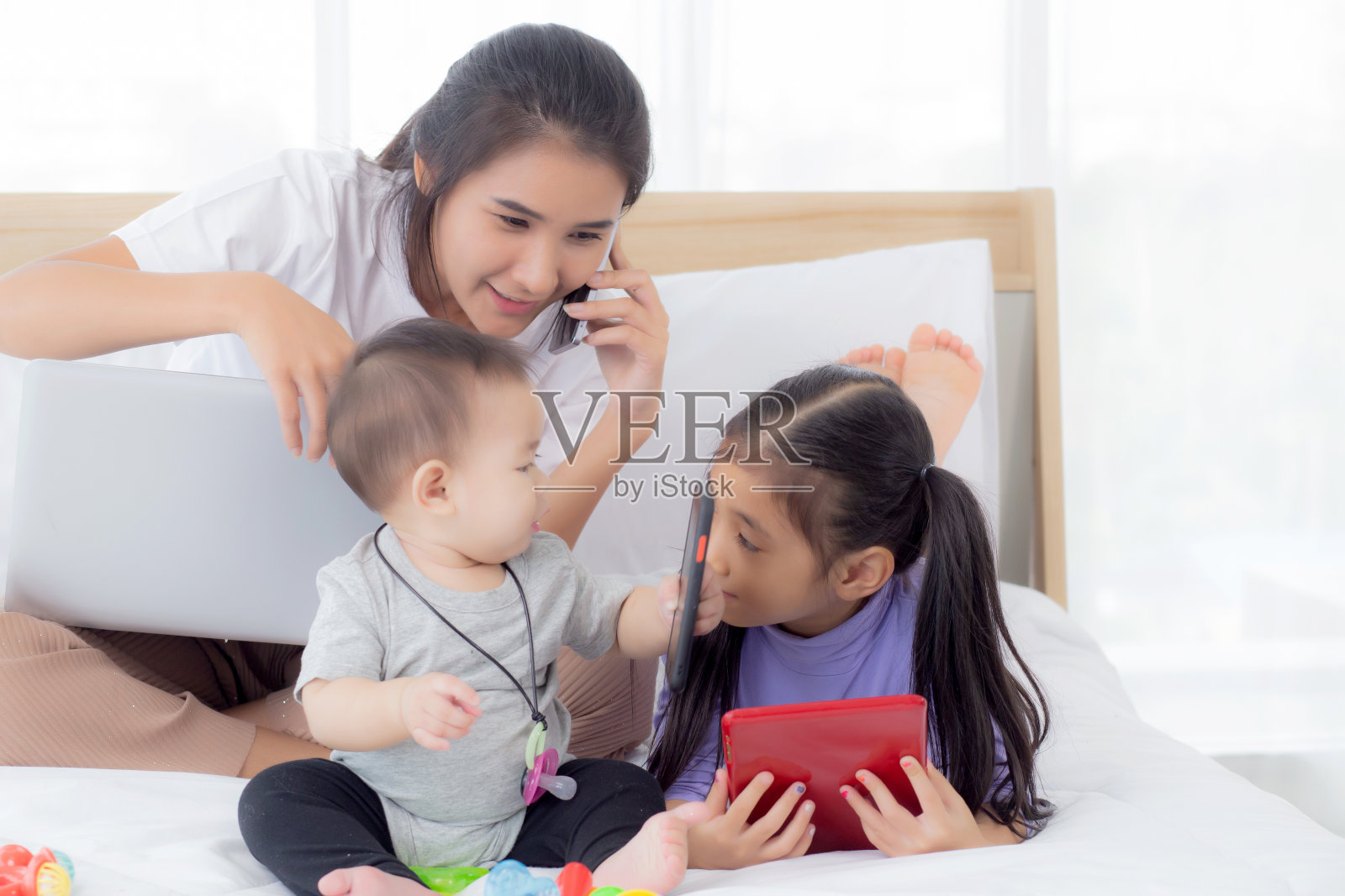 年轻的亚洲母亲在家里工作上网与笔记本电脑和通话电话和女儿看平板电脑在床上卧室，商业女性是自由职业者，新常态，家庭的概念。照片摄影图片