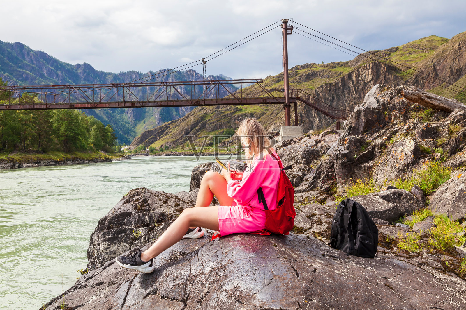 一位年轻漂亮的金发女郎，身穿粉色套装和短裤，坐在阿尔泰山河边的一块大石头上，以一座古老的铁桥为背景看着手机。照片摄影图片