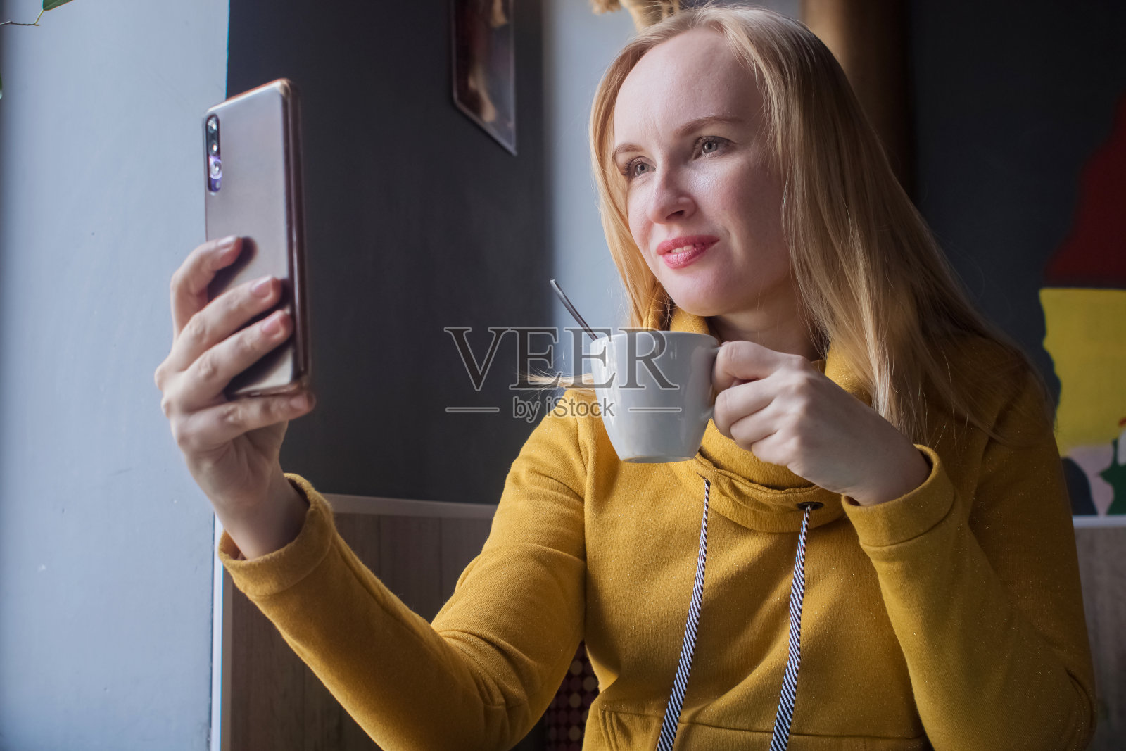 博主女孩坐在咖啡馆里用智能手机拍一杯卡布奇诺泡沫。在社交网络、视频博客和照片博客中工作。互联网内容创作和食物摄影的概念。照片摄影图片