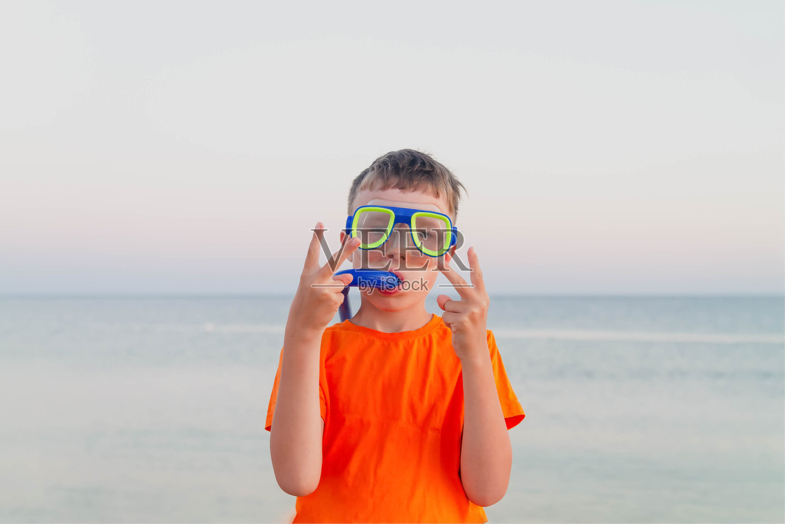 一个戴着水肺面罩和呼吸管的十几岁的男孩。一个穿着橙色t恤的少年在海滩上。那孩子竖起大拇指。那男孩喜欢在海上休息。照片摄影图片