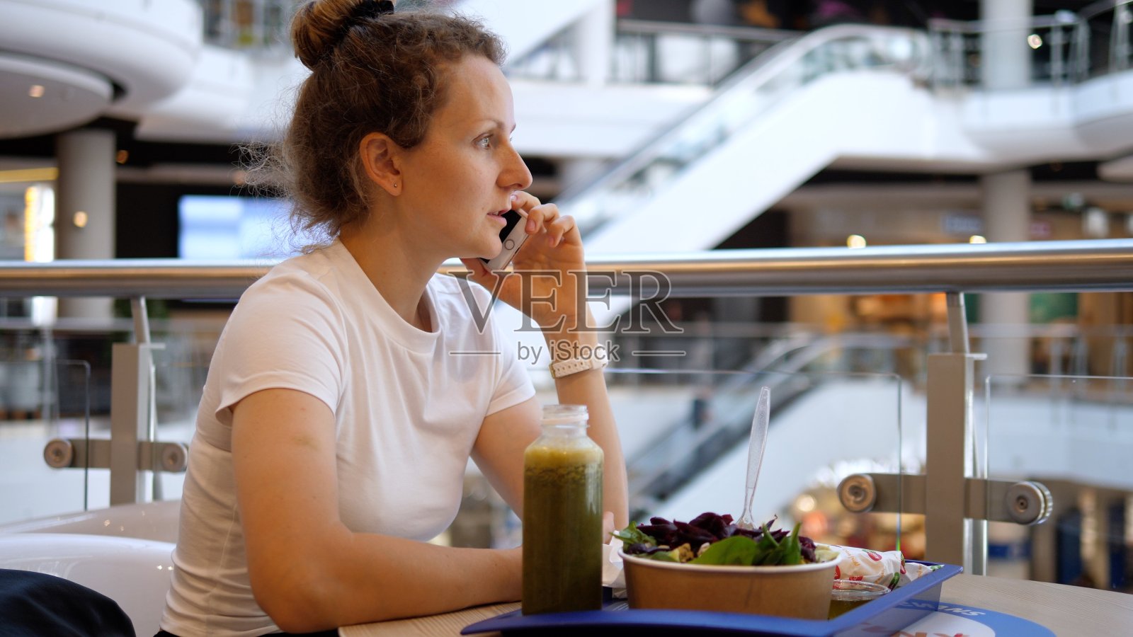 一位年轻女子在购物中心的美食区吃午饭时打电话。绿色奶昔和一份沙拉放在蓝色塑料托盘上照片摄影图片