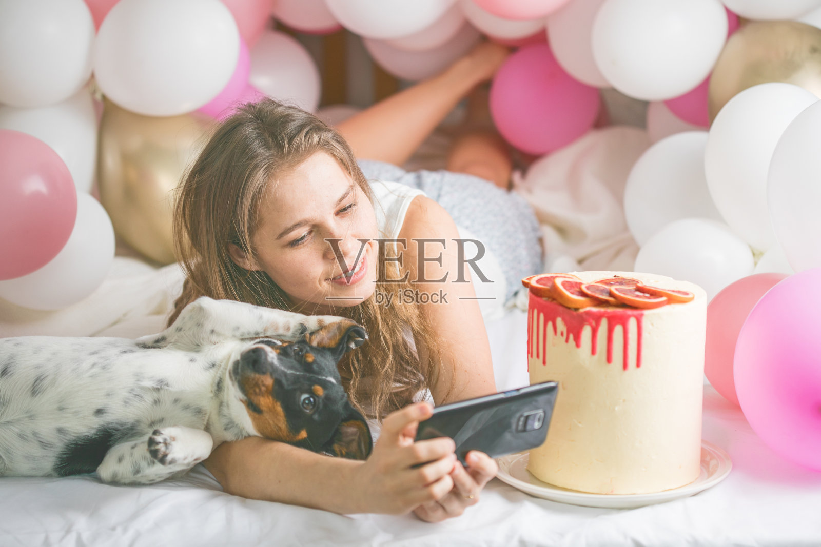 可爱的女士穿着睡衣在卧室里用手机自拍，和她的狗玩。室内肖像女孩在早上与气球。丈夫给妻子惊喜。照片摄影图片