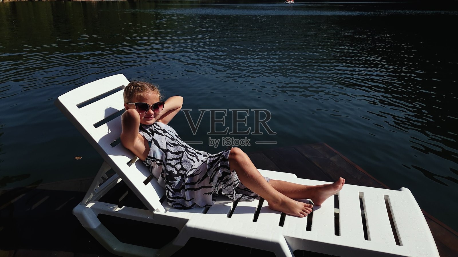 小女孩躺在太阳床上戴着太阳镜，披着波西米亚丝绸披肩。孩子们在水下码头上休息。人行道上覆盖着湖水。照片摄影图片