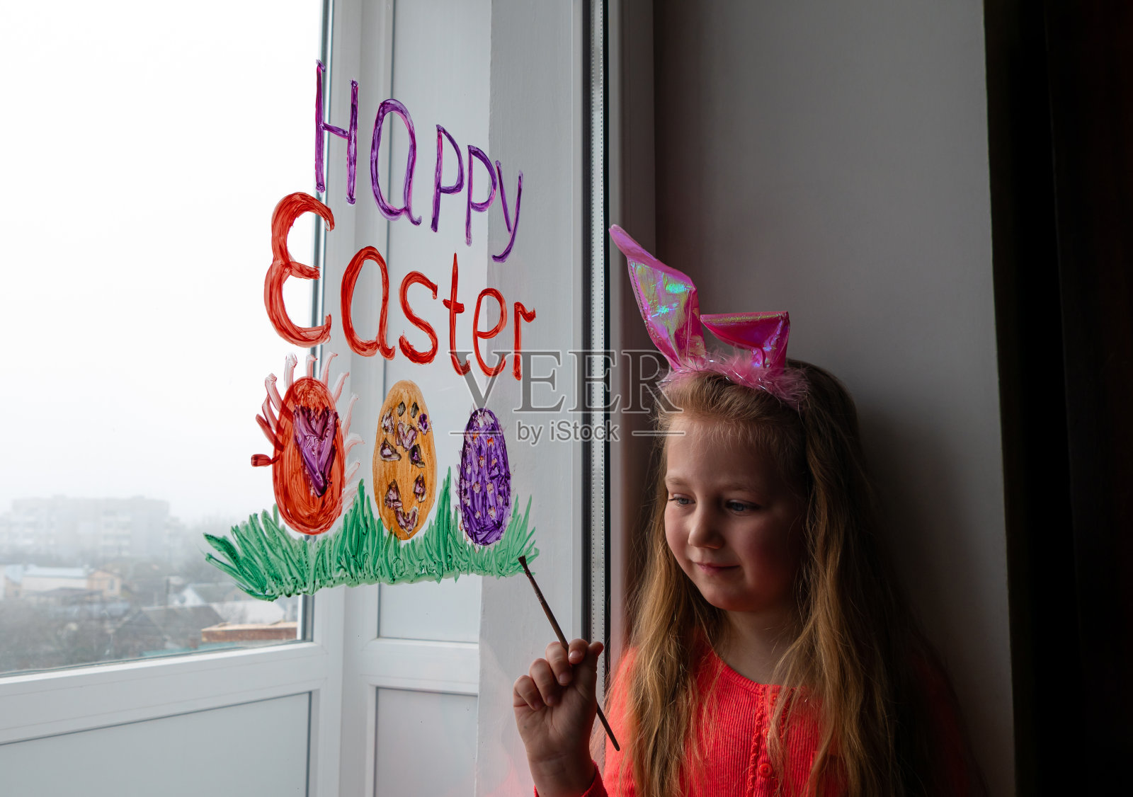 可爱的女孩戴着兔子耳朵在窗户上画复活节彩蛋绿草。孩子准备画节日的家饰照片摄影图片