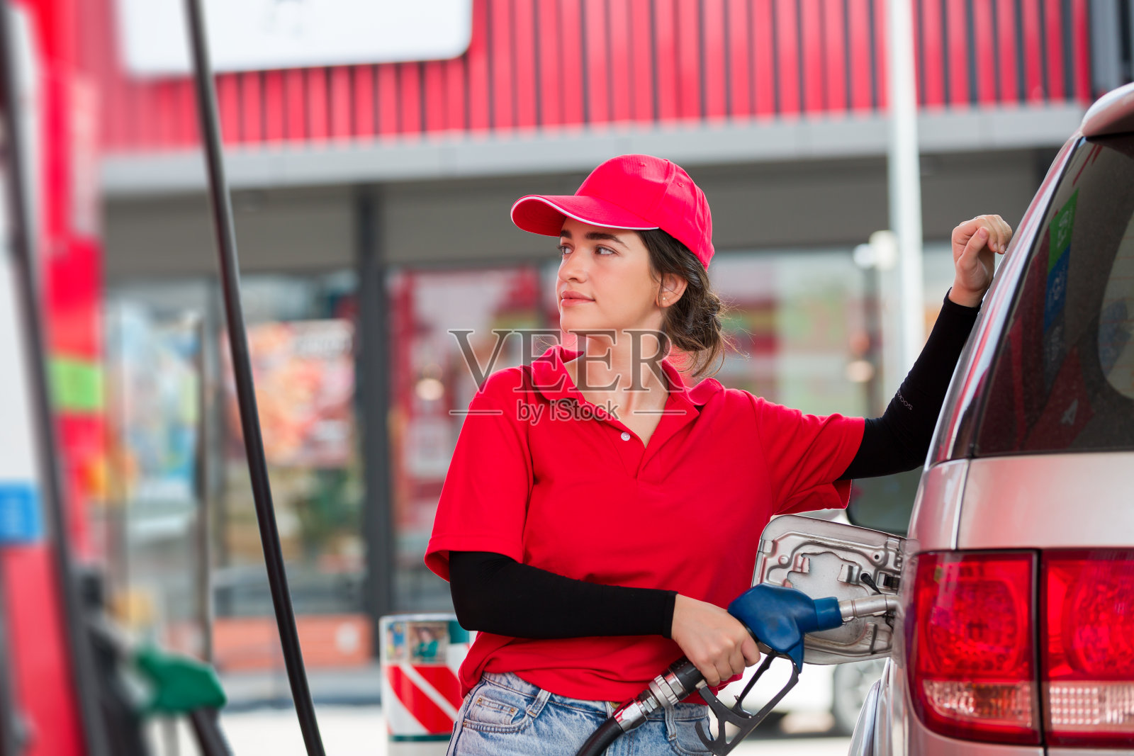 女服务员拿着管嘴在加油站加油。年轻的女助理在加油站加油，笑容满面，穿着红色制服和红帽子照片摄影图片