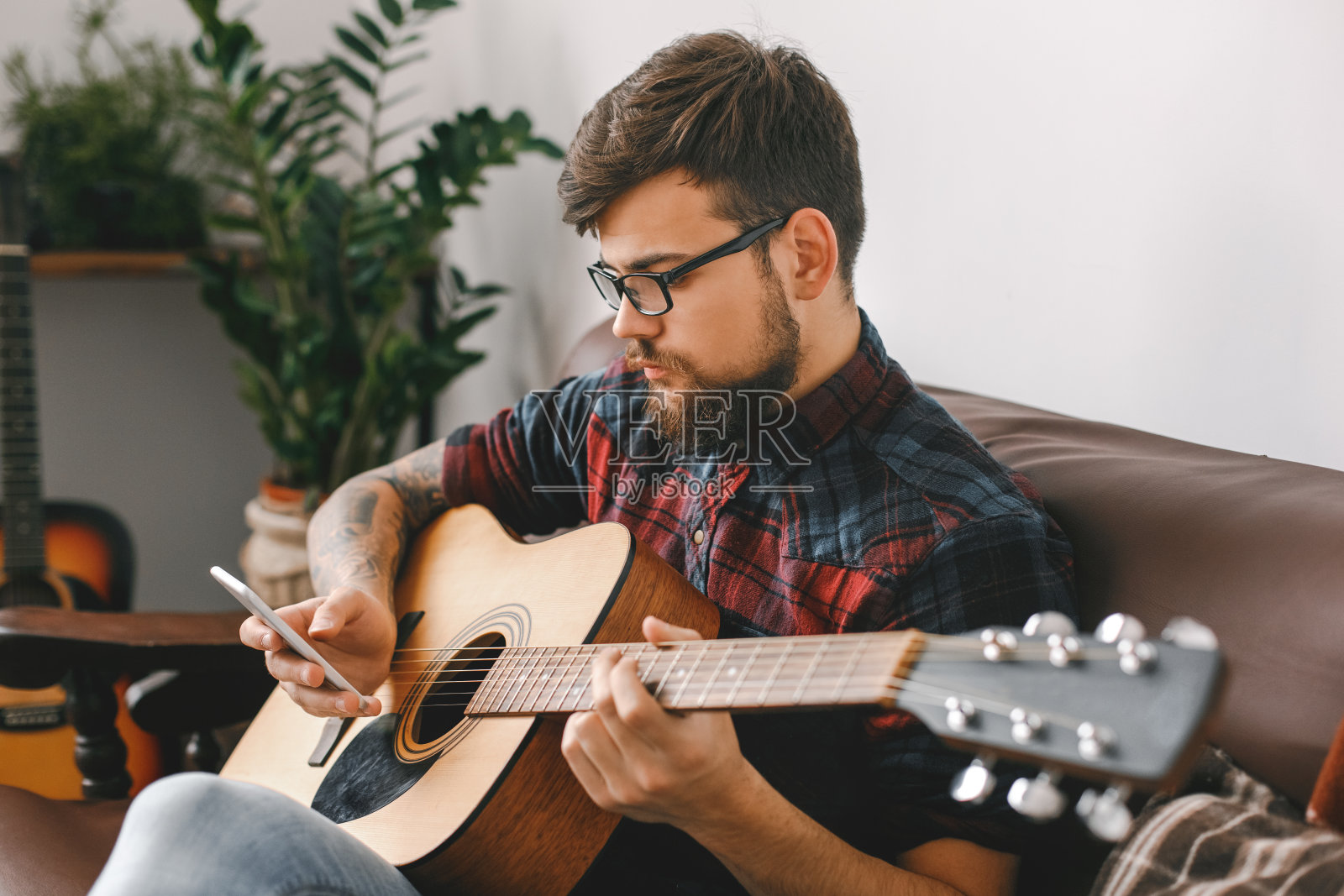 年轻的吉他手潮人在家拿着吉他坐着浏览智能手机检查媒体照片摄影图片