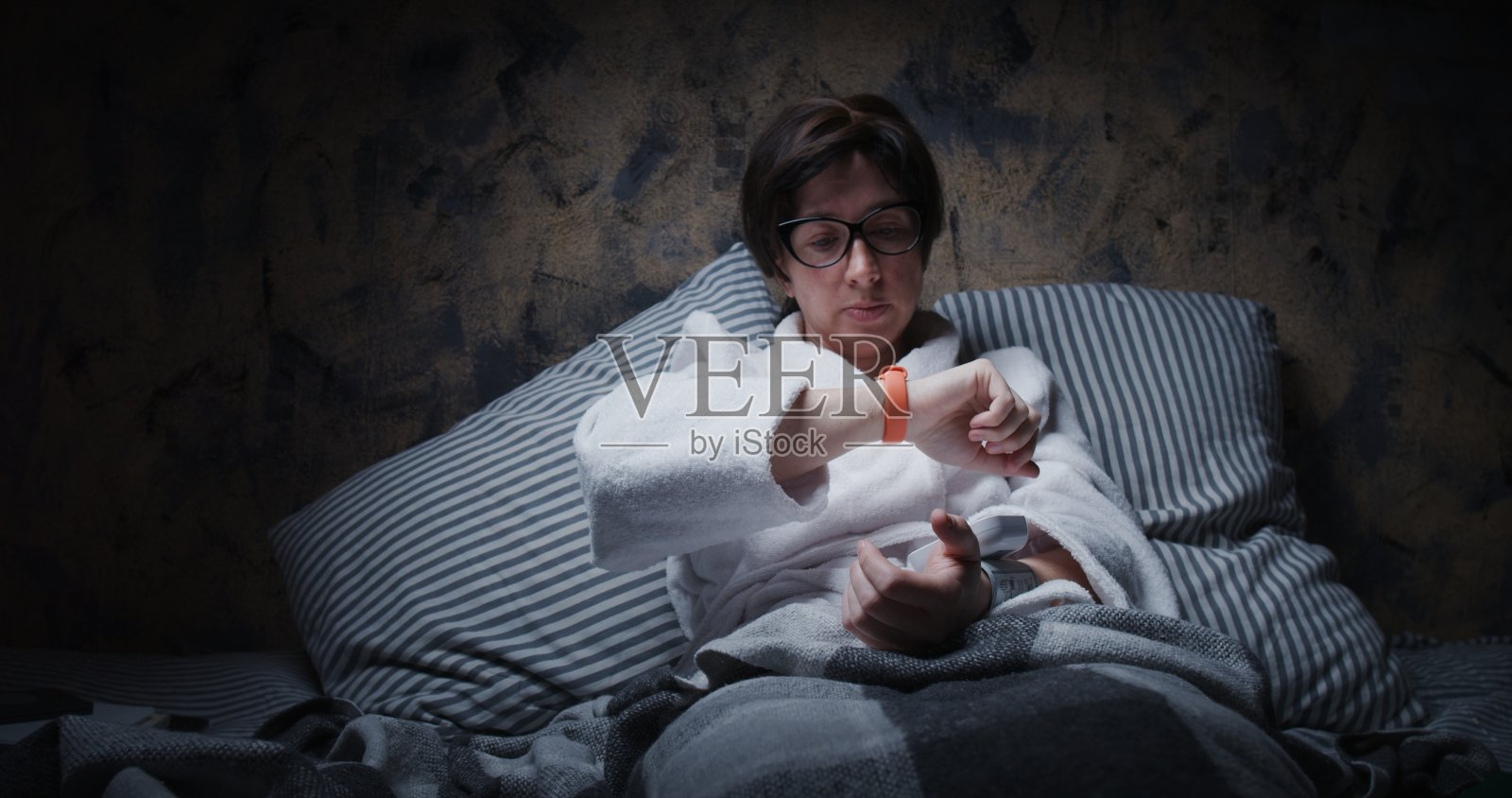 成熟的女人坐在床上，用腕表和智能腕带测量她的血压照片摄影图片