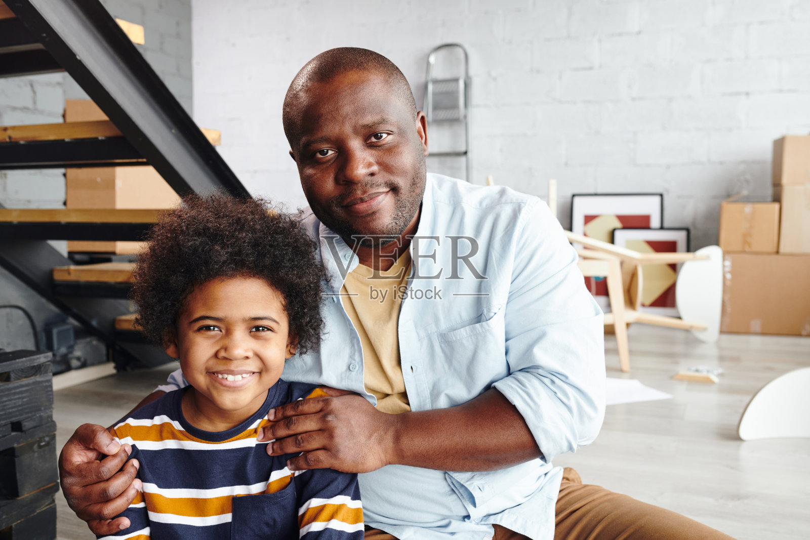 快乐的非洲人拥抱他的可爱的儿子在条纹套衫在起居室的楼梯照片摄影图片