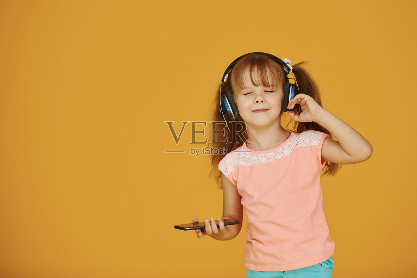 一个带着耳机和手机的可爱小女孩的肖像在录音室里，背景是黄色的照片摄影图片