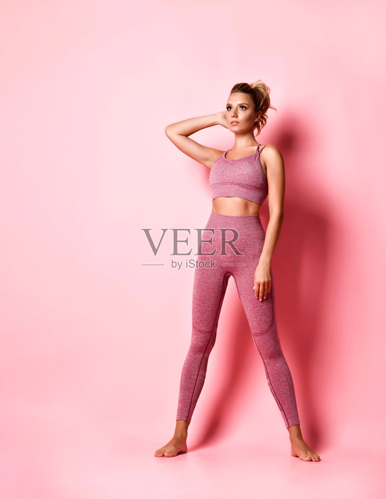 苗条可爱的女士在时尚的玫瑰顶部和紧身裤站起来把一只手放在后面的头。完整的长度肖像孤立在粉红色。本空间照片摄影图片