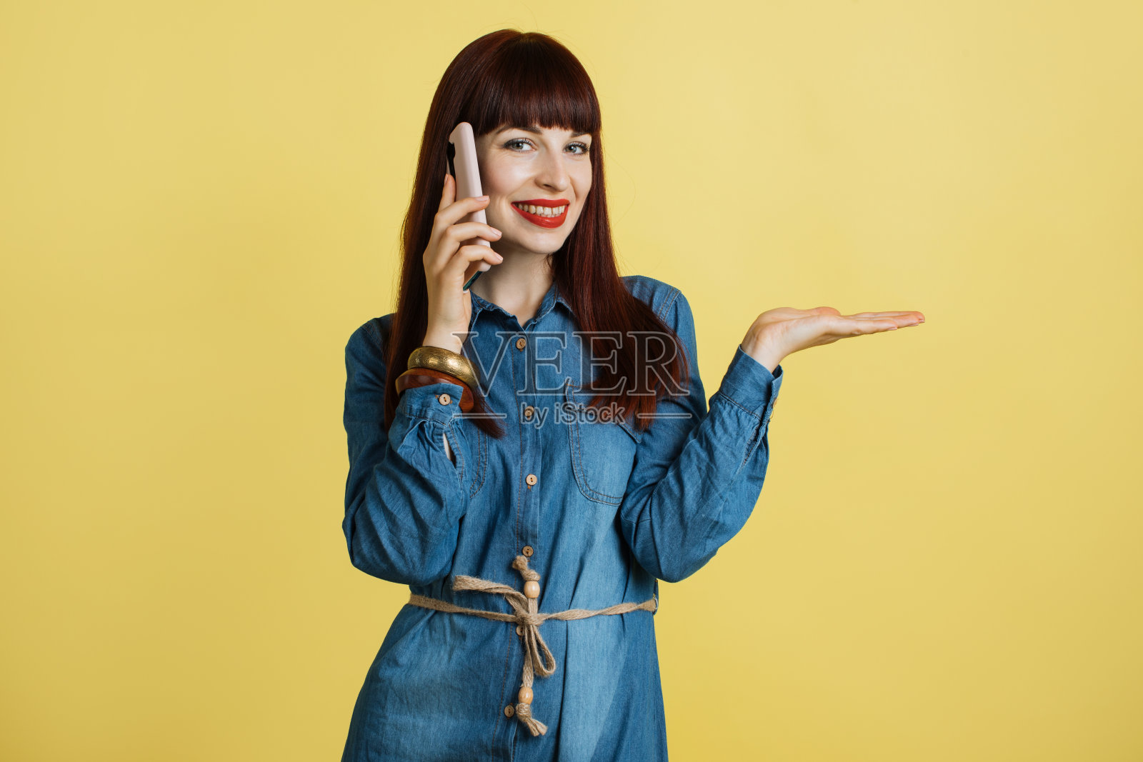 穿着牛仔裤的漂亮女孩的肖像，正在讲电话，显示想象的东西，一些文字，产品或口号的空白复制空间，在张开的手掌上，孤立在黄色的背景。照片摄影图片