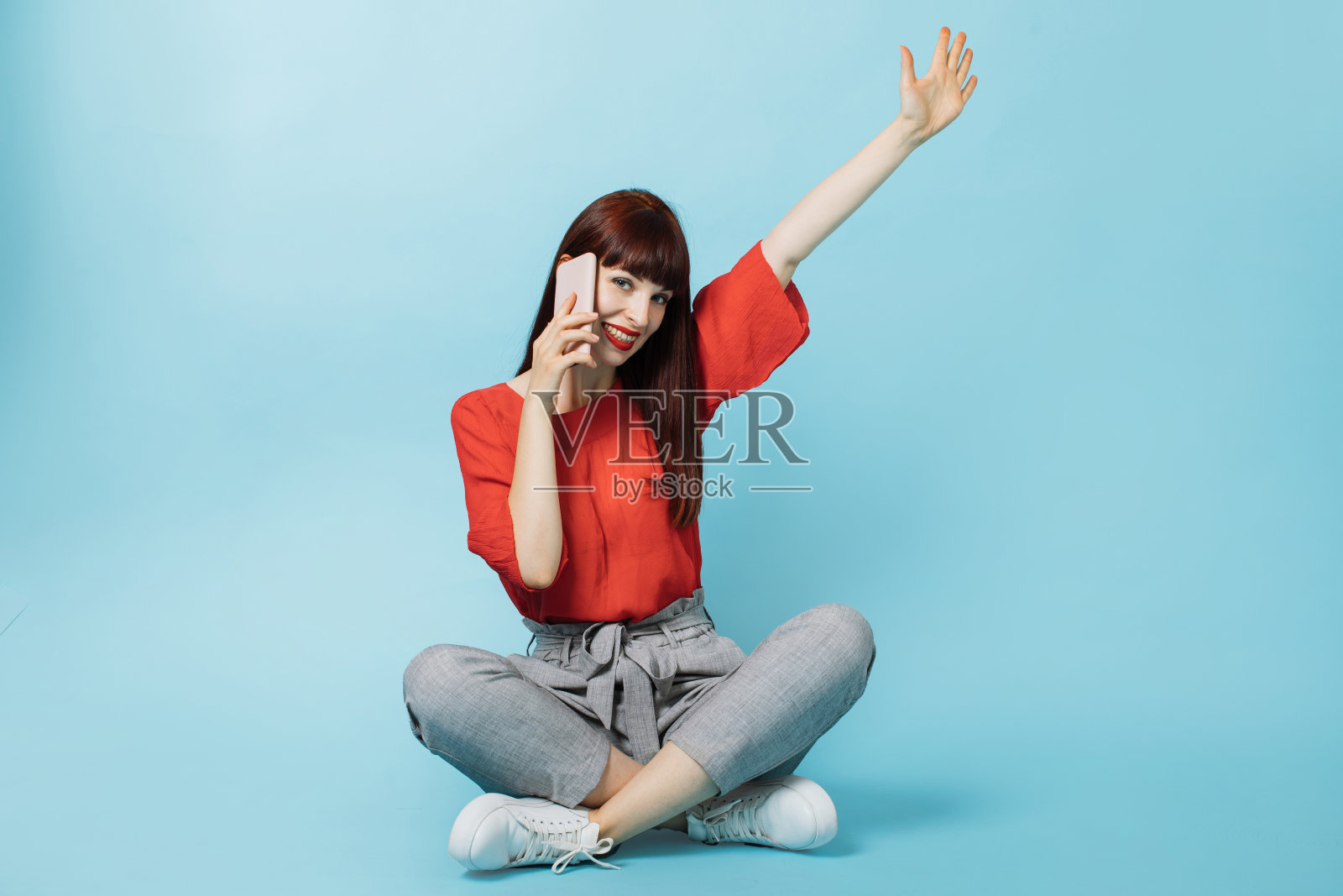 高兴愉快的女人，长红直发，穿着鲜艳的正式衣服，坐在荷花姿势，举起一只胳膊，在孤立的蓝色背景，讲手机照片摄影图片