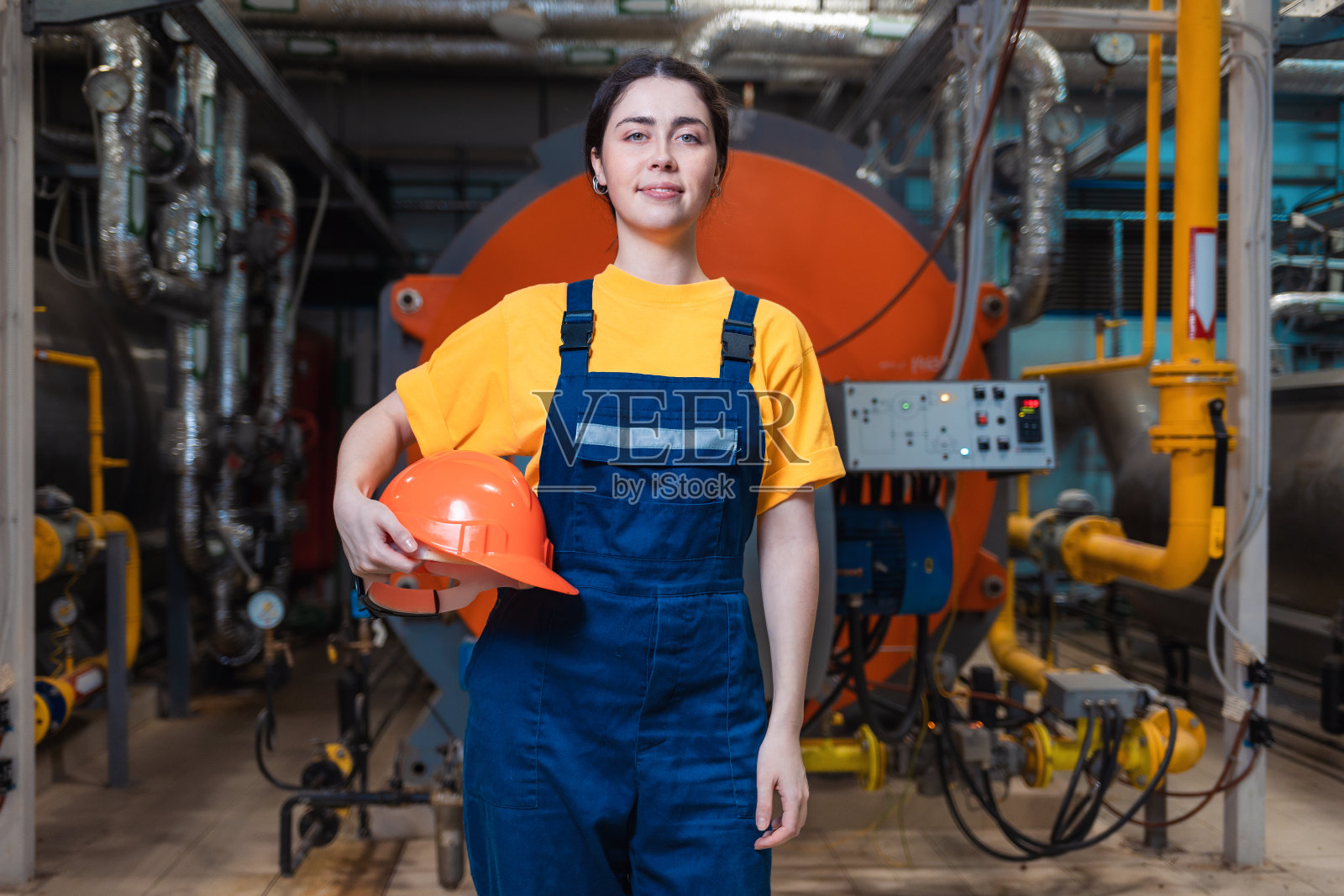 一名身穿制服、面带微笑的年轻女工人，手里拿着安全帽，站在锅炉房里摆姿势。工业生产的概念照片摄影图片